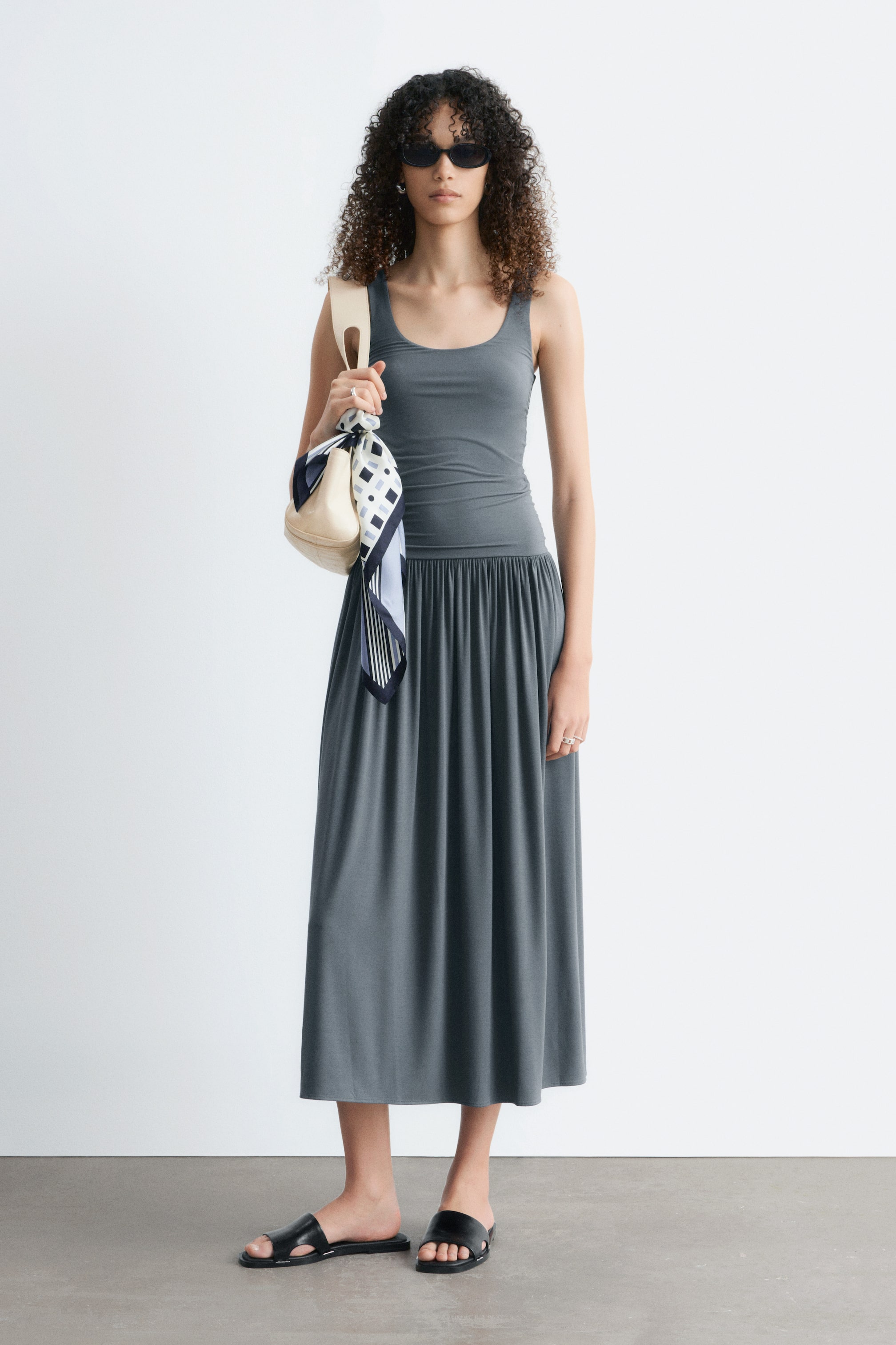 Grotere afbeelding bekijken: Een donkergrijze mouwloze jurk met een aansluitend lijfje met ronde hals dat in de taille is getailleerd met plooien en overgaat in een rok tot op midi-lengte.