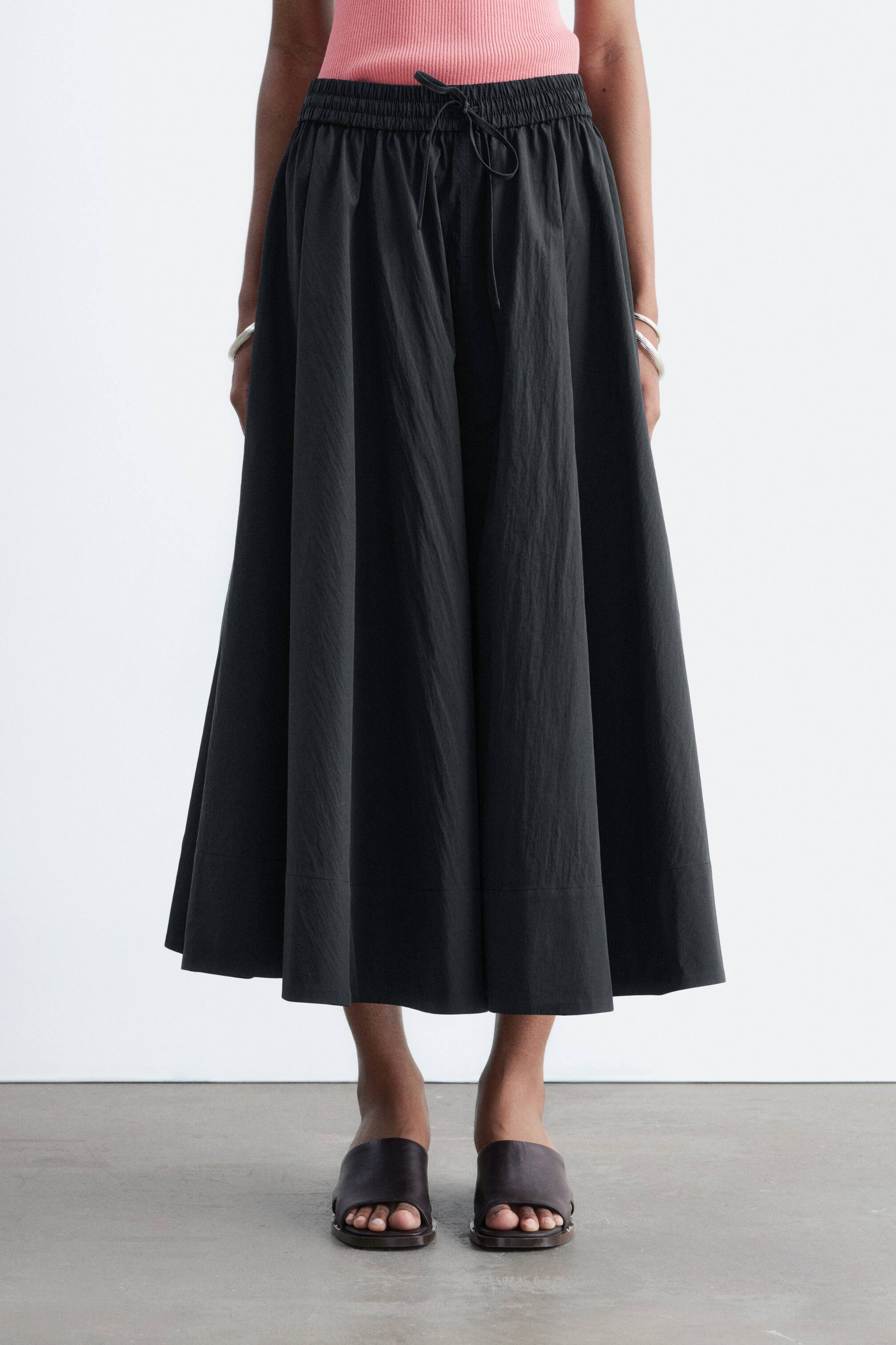 View larger image: The model is wearing a dark, gathered maxi skirt with an elasticated waistband and tie. She is also wearing dark leather slip-on sandals with a wide strap and a thick sole.