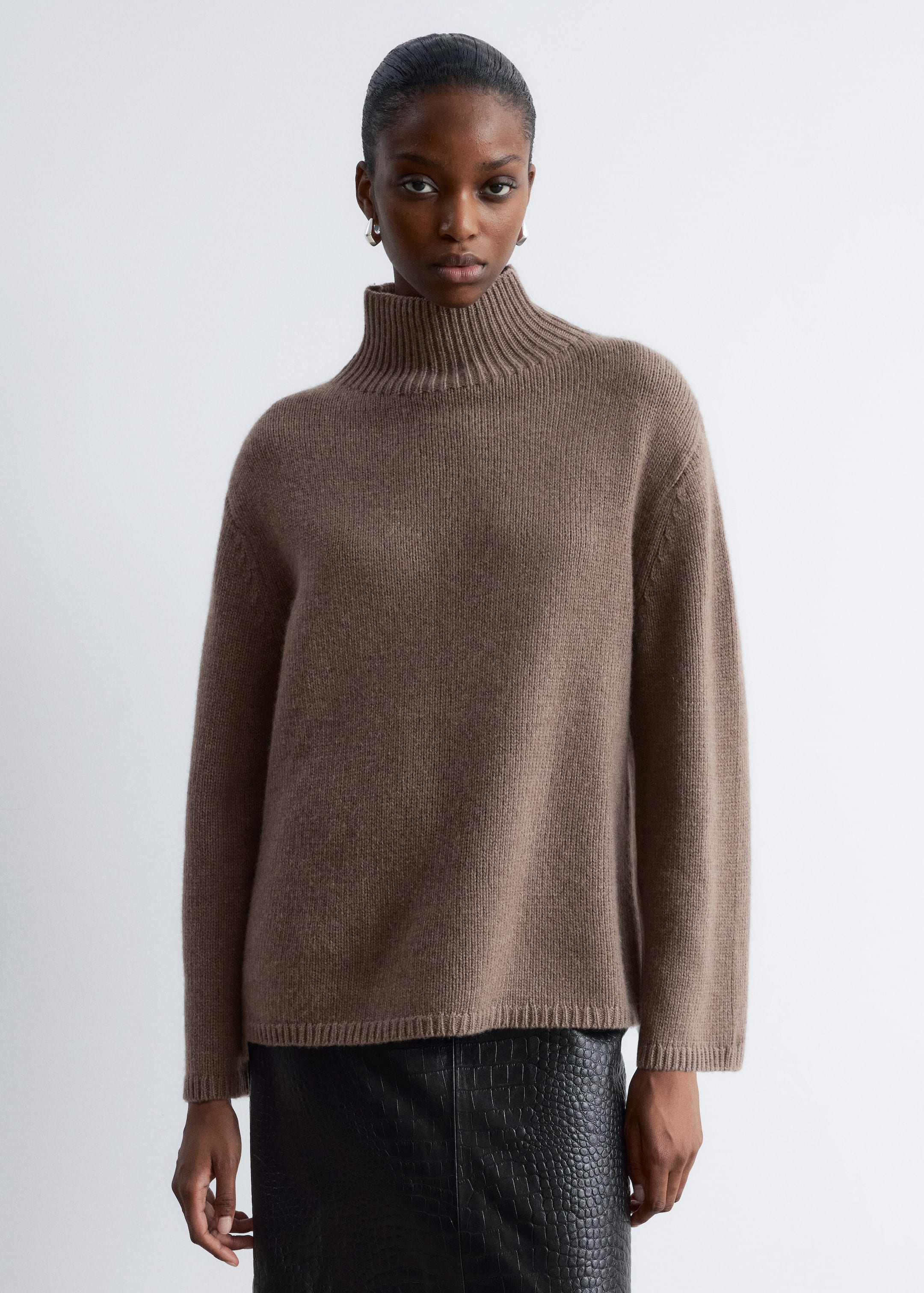 Grösseres Bild ansehen: Ein taupefarbener Rollkragenpullover wird getragen, der eine lockere Passform, überschnittene Schultern und lange Ärmel mit gerippten Bündchen aufweist. Er präsentiert einen hohen, gerippten Rollkragen und einen gerippten Saum, der über einen schwarzen, geprägten Rock reicht.