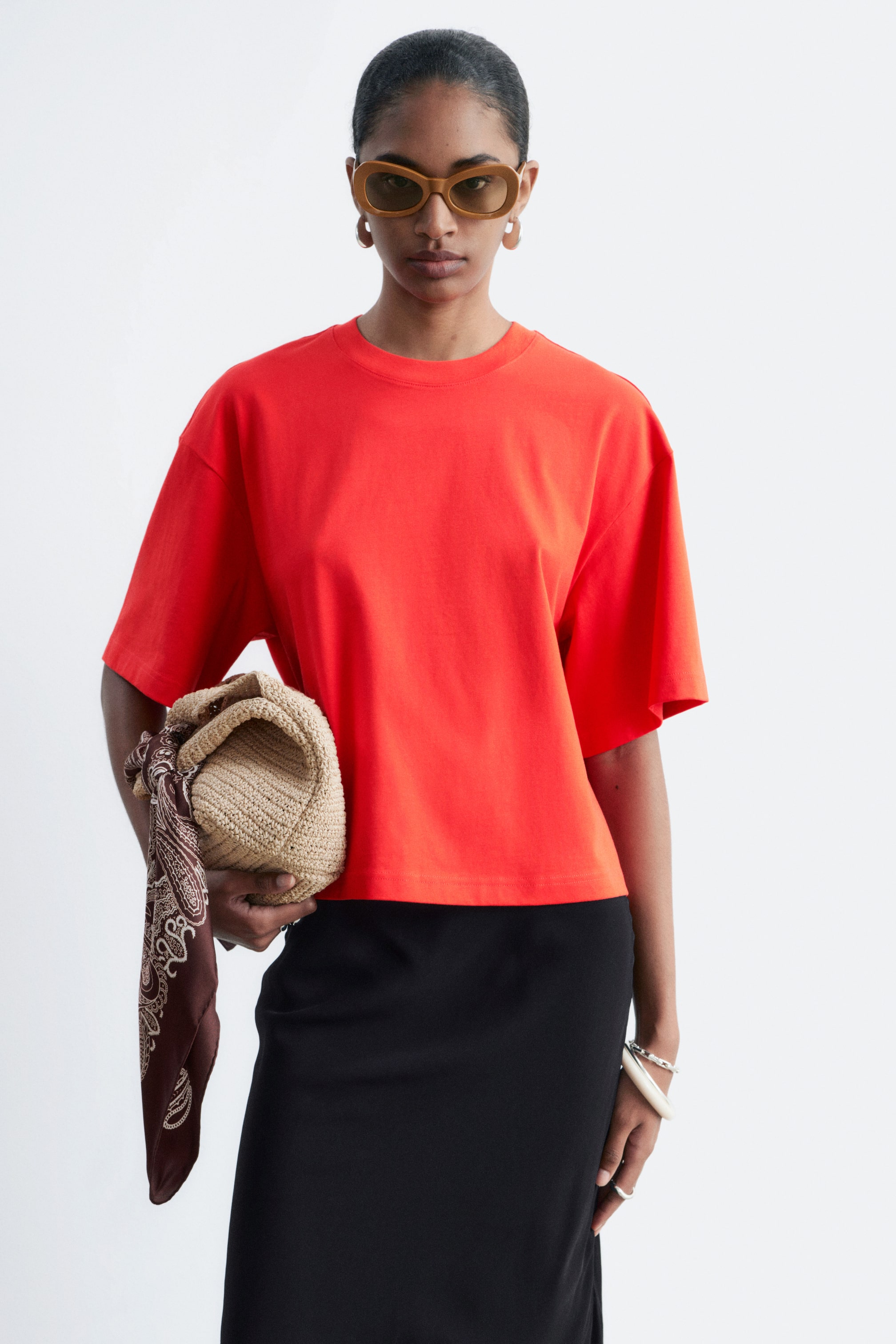 Ver imagen más grande: La modelo lleva una camiseta roja con cuello redondo clásico y mangas cortas y anchas. La camiseta presenta un ajuste relajado y cuadrado, con un dobladillo recortado, y está combinada con una falda negra.