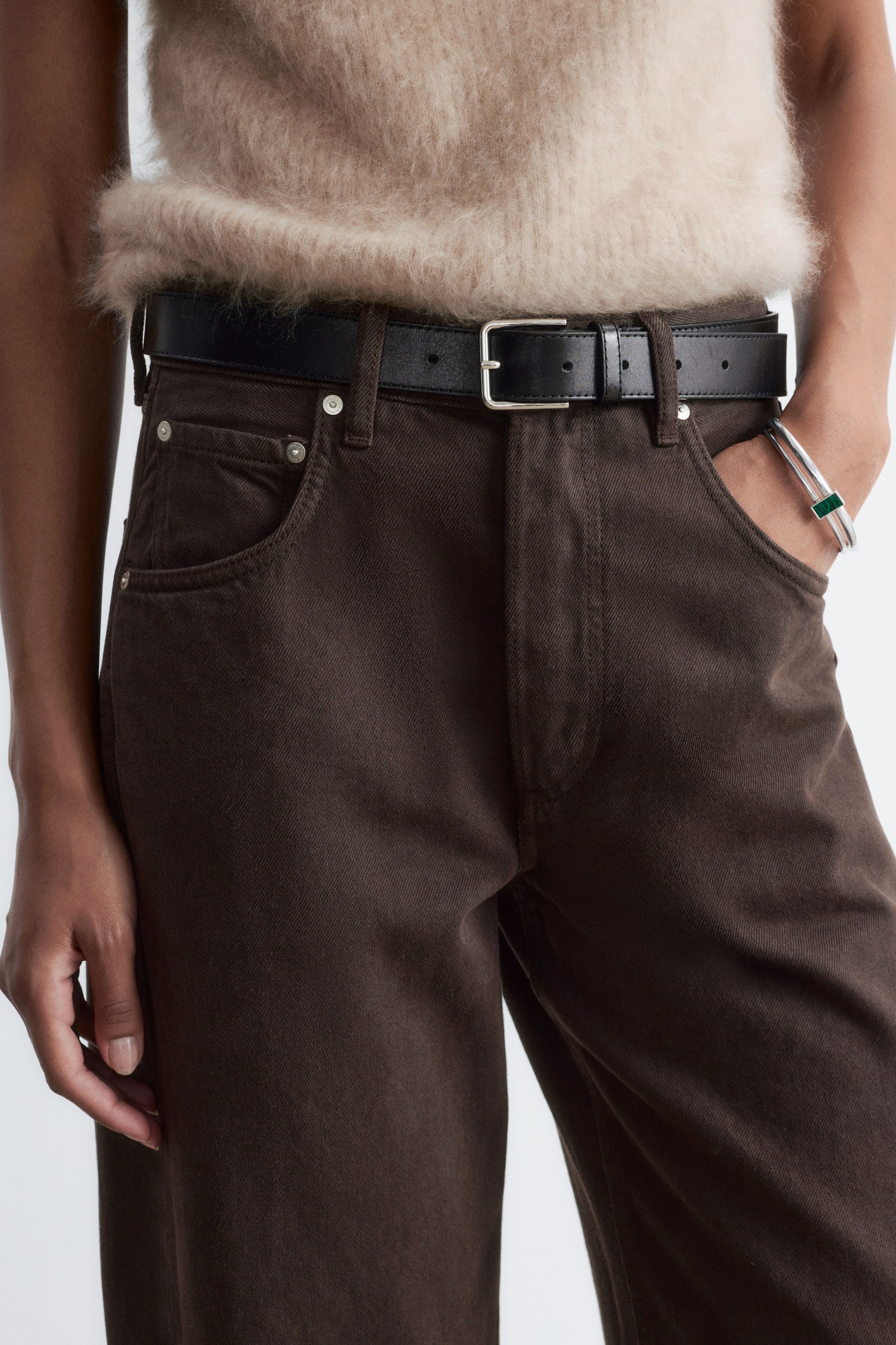 Ver imagem maior: Jeans castanho-escuros, de cintura média, com um design clássico de cinco bolsos, usados pela modelo com um cinto de couro preto e um top bege de textura felpuda por dentro das calças. A modelo está de pé, com uma mão colocada no bolso da frente.