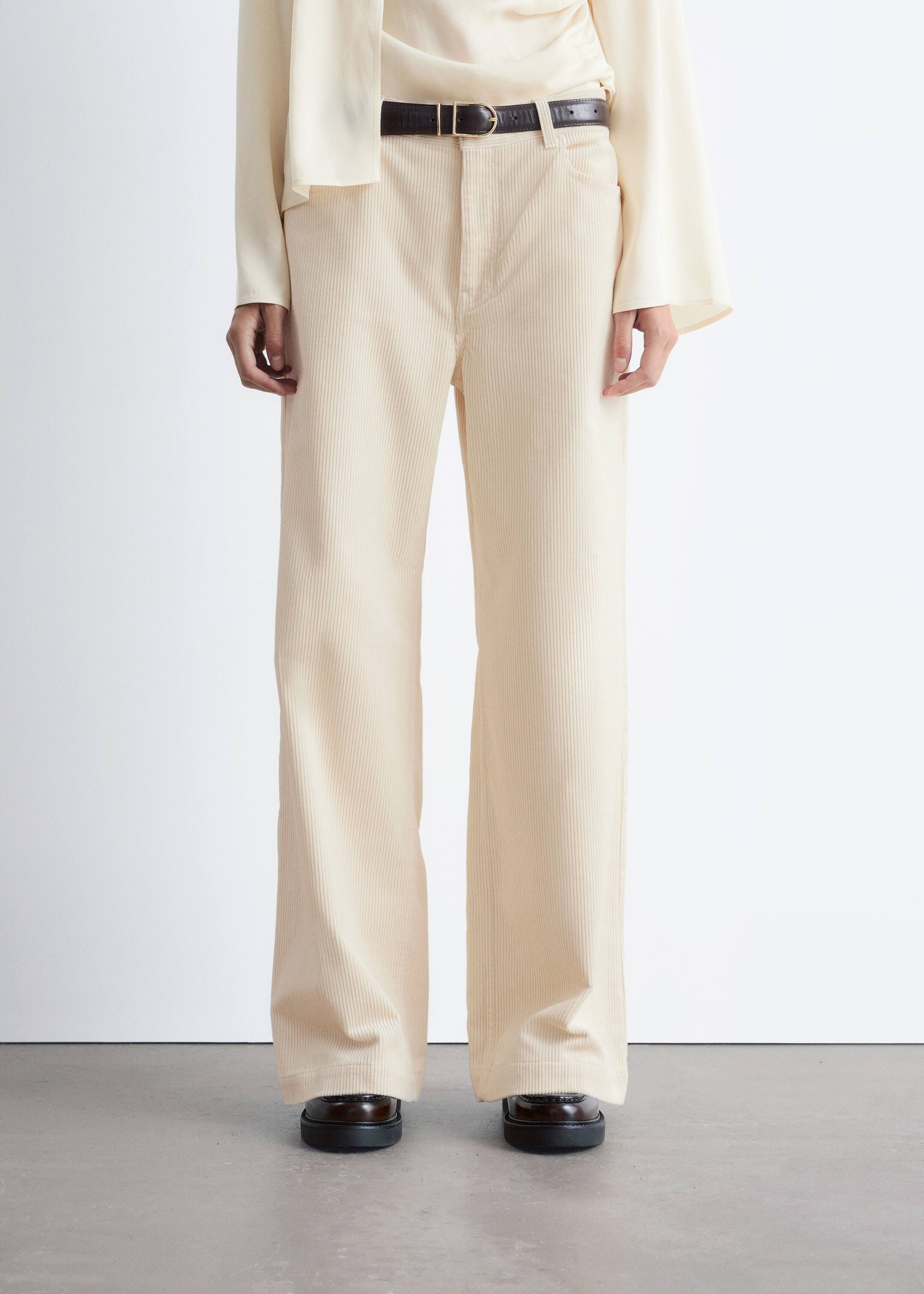 Visualizza immagine più grande: I pantaloni di velluto a coste color crema sono mostrati frontalmente, caratterizzati da una silhouette a gamba larga e una vestibilità a vita alta. I pantaloni sono abbinati a una cintura scura attraverso i passanti e si accumulano leggermente sopra le scarpe scure, indicando la loro lunghezza completa.