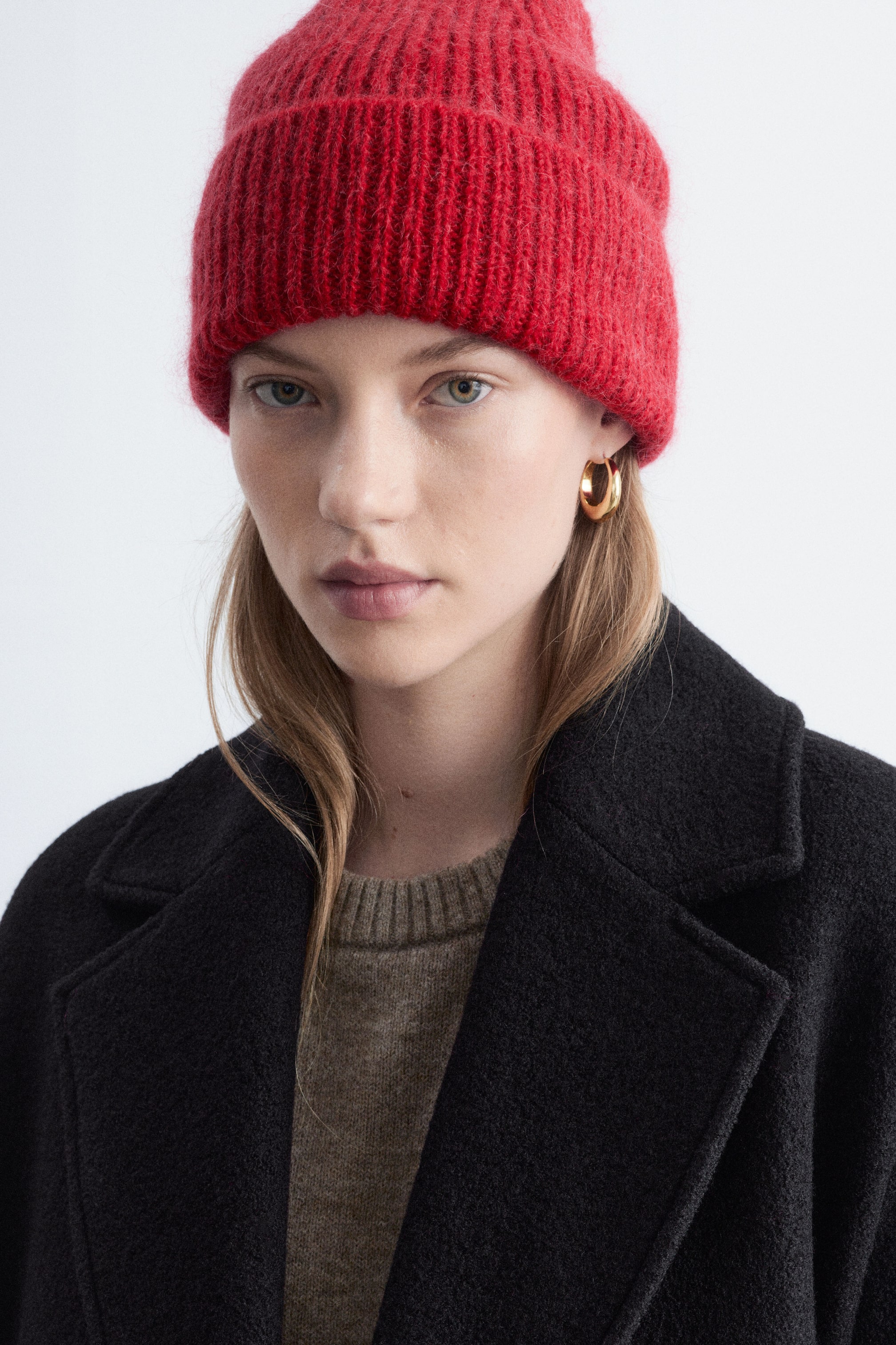 View larger image: A red rib-knit beanie with a fuzzy, textured finish and a wide folded brim is being worn by the model. The beanie is styled with a black coat and a beige crew neck sweater.