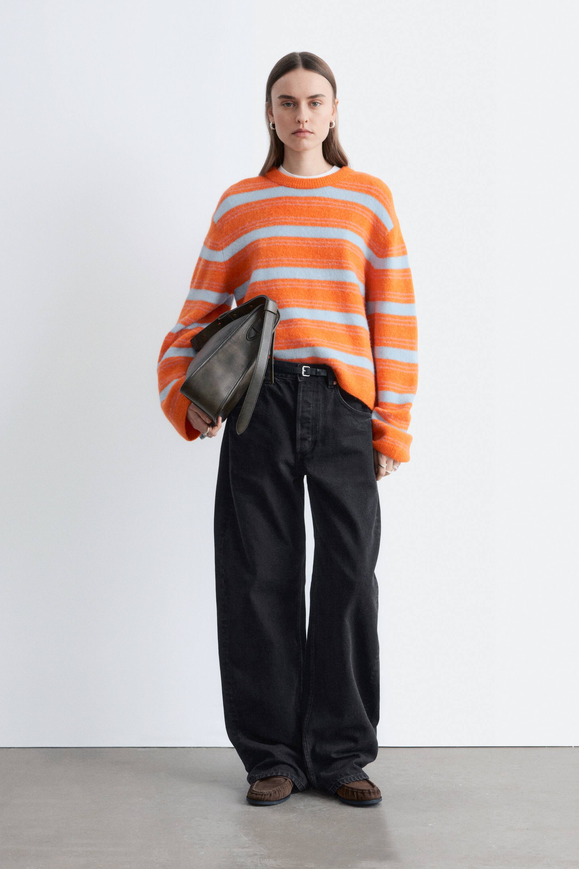Преглед на по-голямо изображение: Оверсайз, пухкав пуловер с оранжеви и светлосини райета и кръгло деколте, с рипсени маншети, е облечен върху бяла тениска. Комбиниран е с широки черни дънки с широки крачоли, стегнати с черен колан със сребърна тока. Тъмно маслинено-зелена чанта през рамо е държана в сгъвката на лявата ръка, а кафяви мокасини в стил лоуфър завършват тоалета.