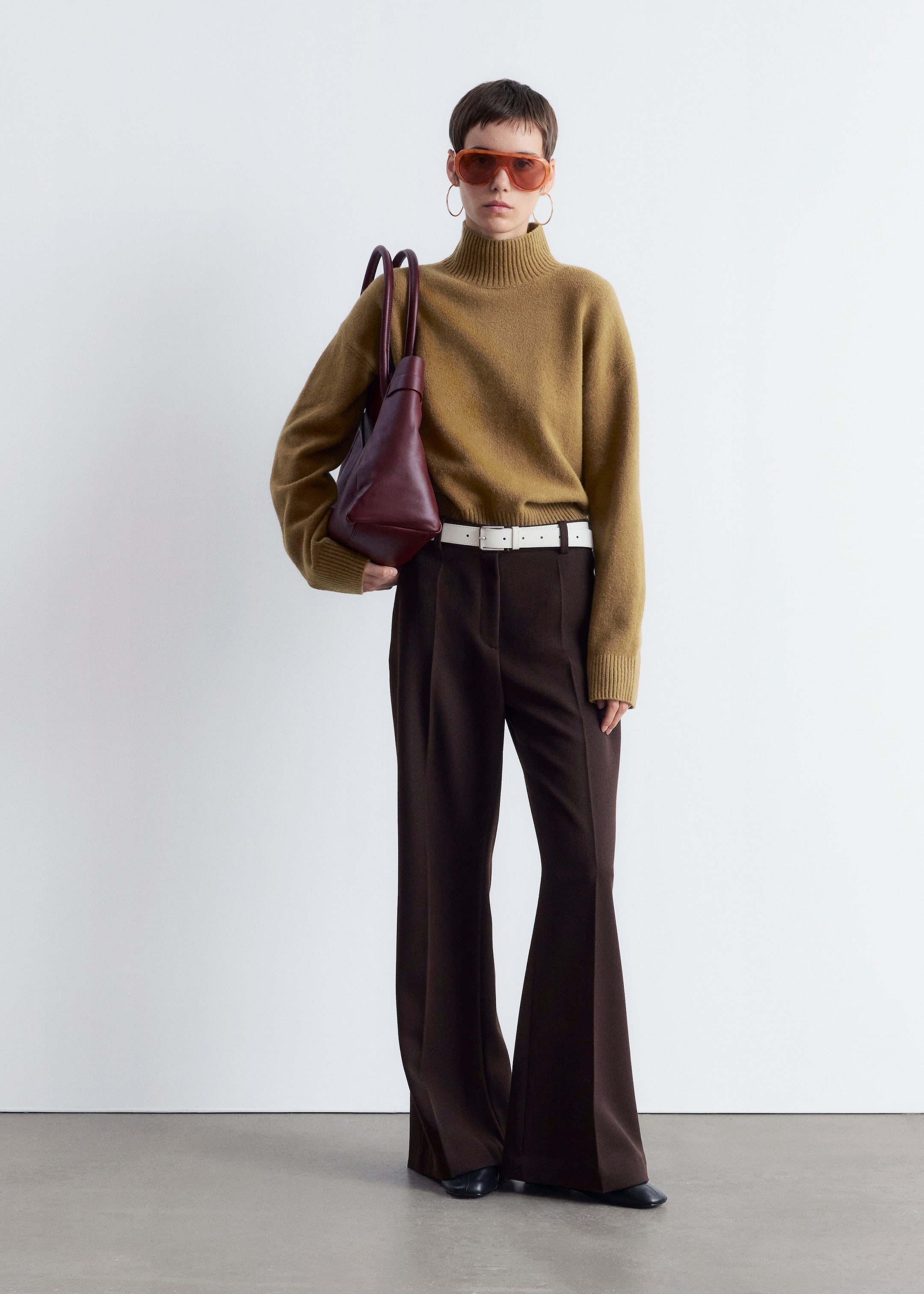 Agrandir l'image: Une personne présente une tenue composée d'un pull à col montant couleur moutarde avec des poignets et un ourlet côtelés, assorti à un pantalon large marron foncé et une ceinture blanche. Ils portent un sac structuré à poignée supérieure rouge foncé sur l'épaule et portent des chaussures plates à bout carré foncé et des lunettes de soleil surdimensionnées teintées orange.