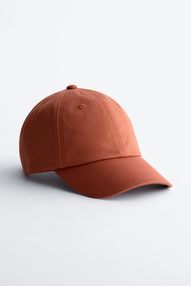 Een honkbalpet vervaardigd uit katoenen keperstof toont zijn voor- en zijkant. Het heeft een roestkleur, een klassieke gebogen rand met sierstiksel, een paneelontwerp en twee ventilatiegaatjes op de zichtbare panelen. - 1