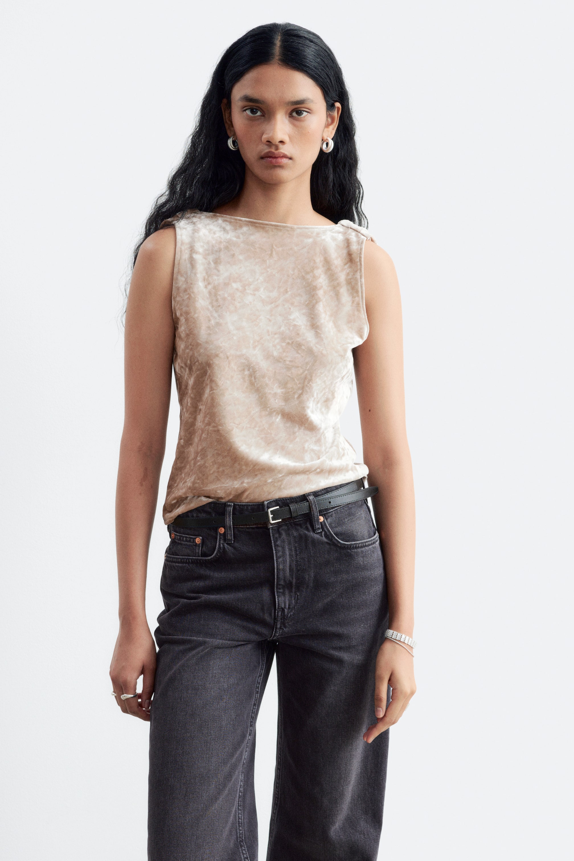 Grösseres Bild ansehen: Ein champagnerfarbenes ärmelloses Oberteil aus Crush-Samt wird vom Model getragen und verfügt über einen geraden U-Boot-Ausschnitt. Das Oberteil ist in dunkelgraue Jeans gesteckt, die mit einem schwarzen Gürtel gehalten werden.