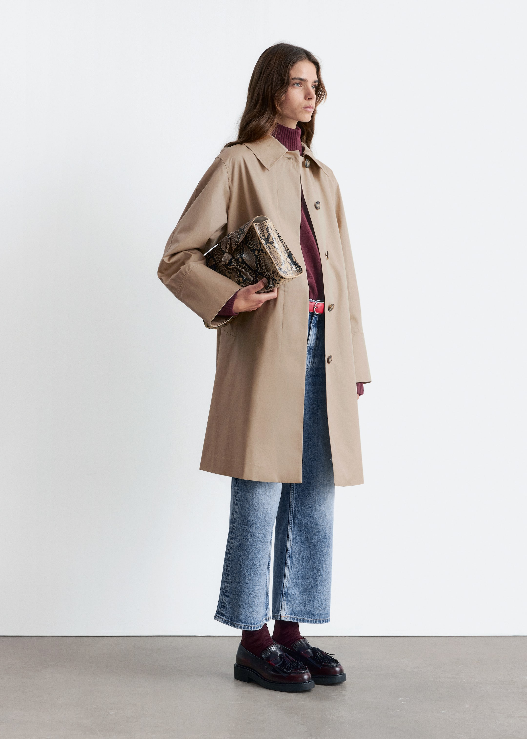 Visa större bild: En person står i en lätt vinkel och visar upp en beige trenchcoat med en klassisk krage och raglanärmar, lager på lager över en vinröd polotröja. Ljusblå, vida, korta jeans syns under kappan, säkrade med ett tunt rött bälte, och kombinerade med mörkröda tofsloafers och vinröda ribbade strumpor, medan en kuvertväska med ormskinnsmönster hålls i ena handen.
