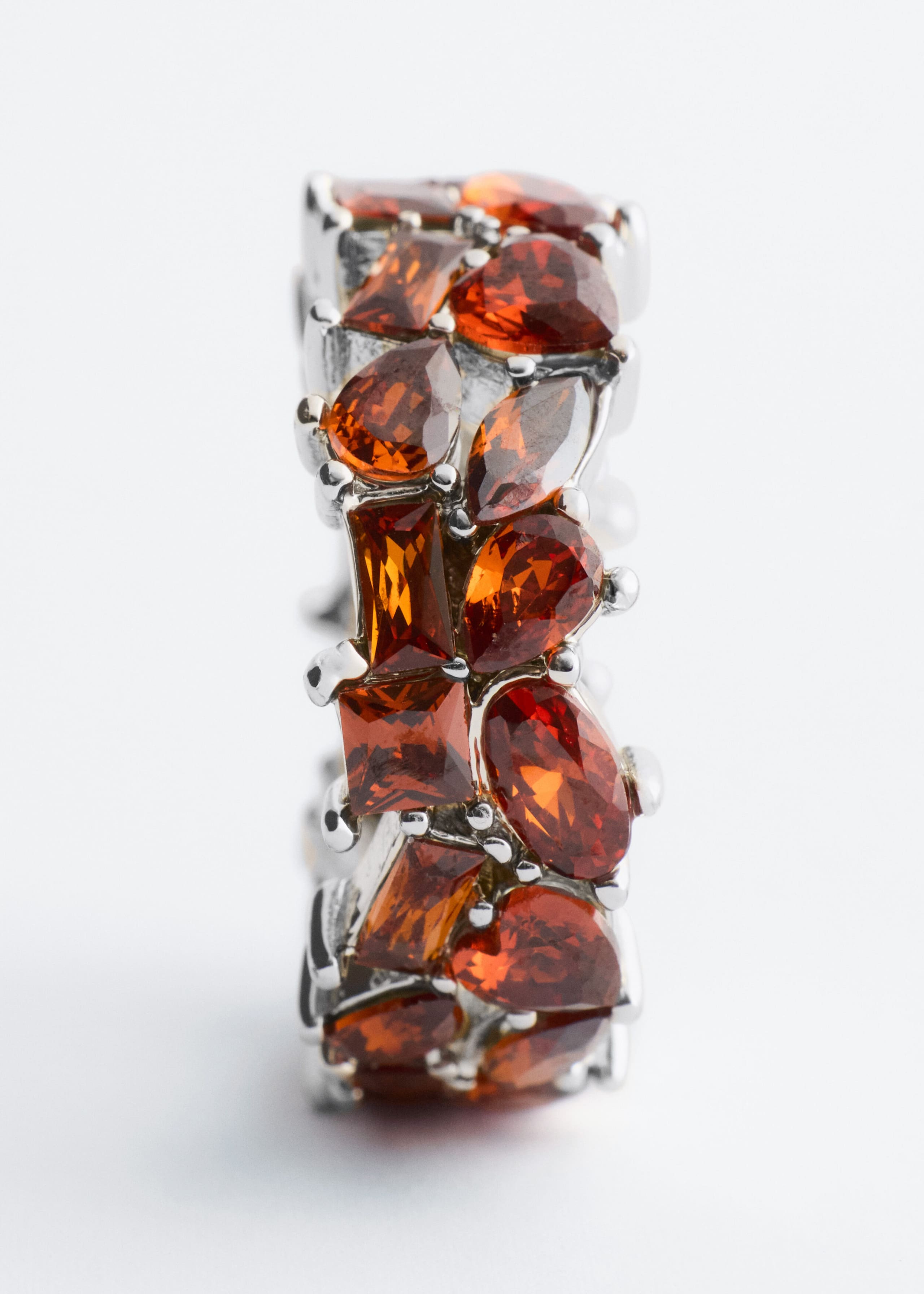 A silver-tone ring is standing vertically, showcasing a front-facing view of its wide band adorned with multi-cut orange and amber gemstones. The gemstones, featuring pear, marquise, oval, and rectangular shapes, are clustering together in a dense pattern secured by small silver-tone prongs.