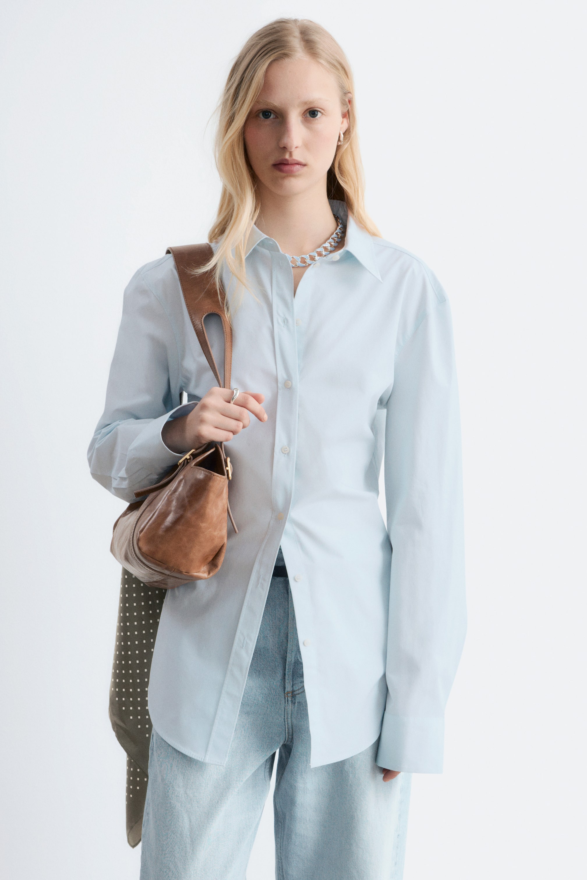 View larger image: A light blue button-up shirt is facing forward, featuring a pointed collar, a front placket with light buttons, and long sleeves with cuffs. It is being worn partially unbuttoned over light-wash jeans, showcasing a silver-toned chunky necklace on the collar and a brown leather bag draping over the left shoulder.