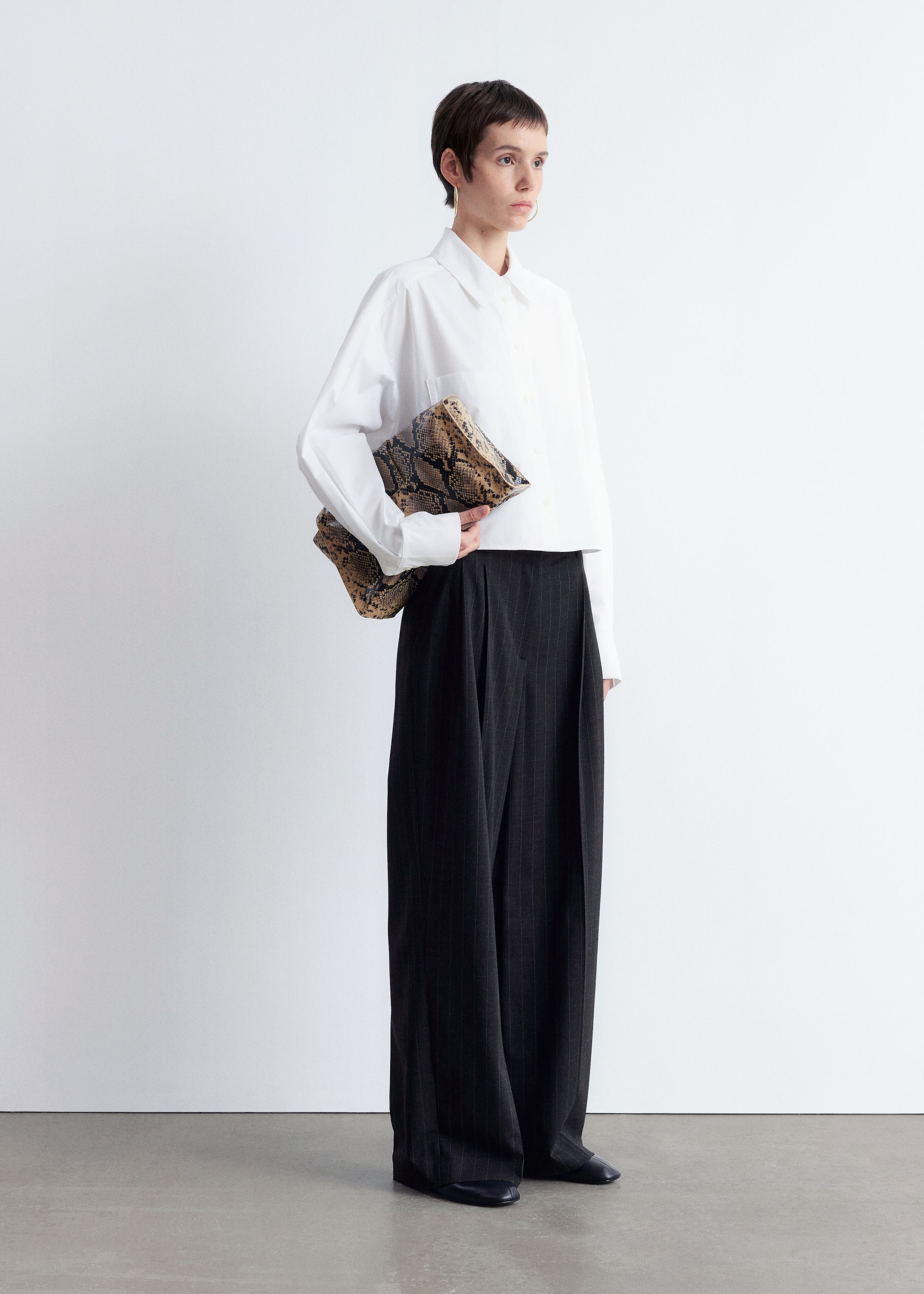 Visualizza immagine più grande: Una camicia bianca oversize con bottoni, caratterizzata da un colletto a punta, una tasca sul petto e maniche lunghe, presenta una vestibilità corta sopra pantaloni neri a gamba larga che mostrano sottili righe e pieghe frontali che si drappeggiano fino al pavimento. Una pochette con motivo a pelle di serpente marrone e nero è tenuta sotto il braccio sinistro.