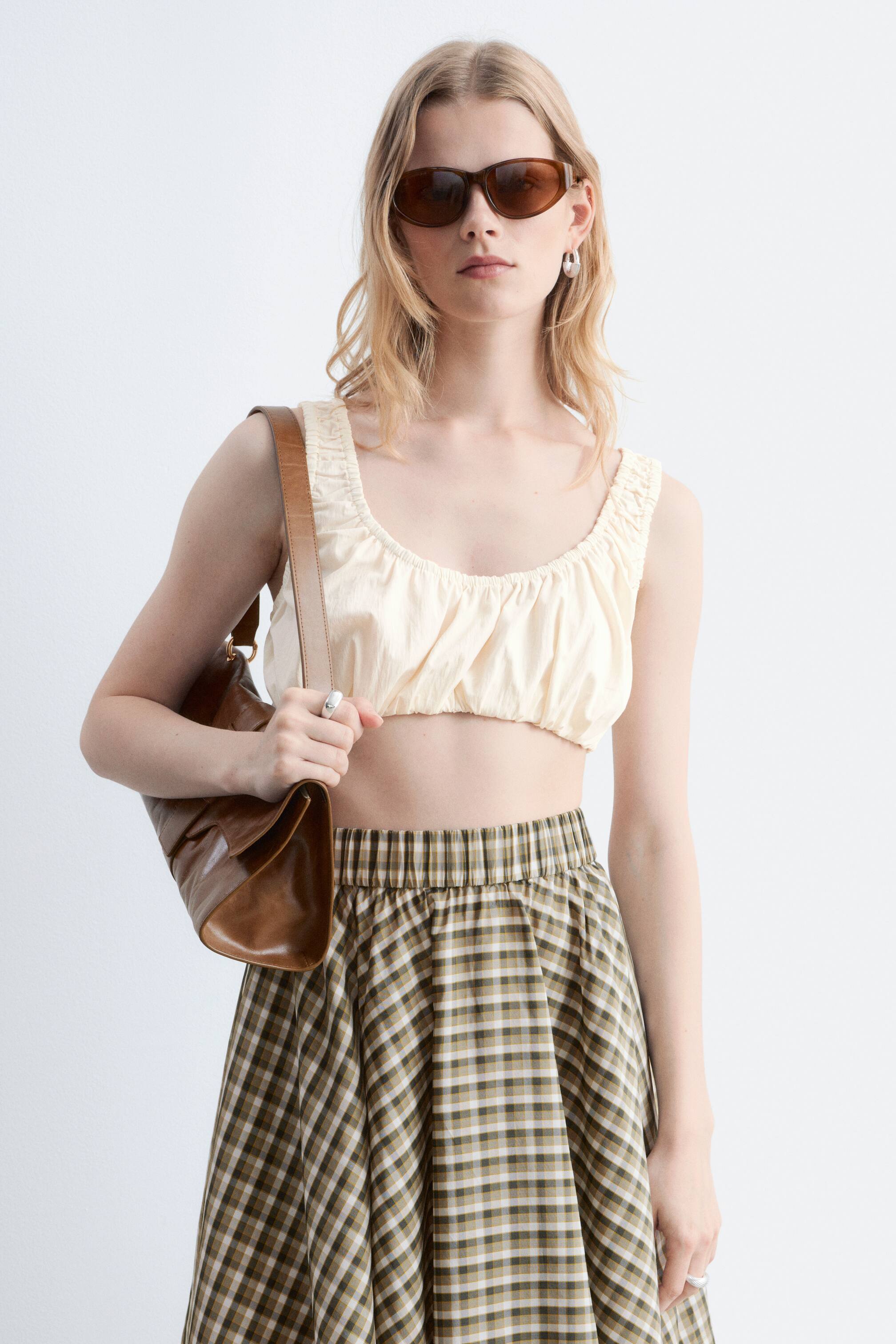 Ver imagem maior: A modelo está a usar um top curto de cor creme com franzidos e decote quadrado, combinado com uma saia midi xadrez e uma mala de ombro em pele castanha.