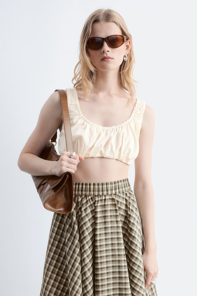 La modelo lleva puesto un top corto de color crema con fruncidos y escote cuadrado, combinado con una falda midi de cuadros y un bolso de hombro de cuero marrón. - 1