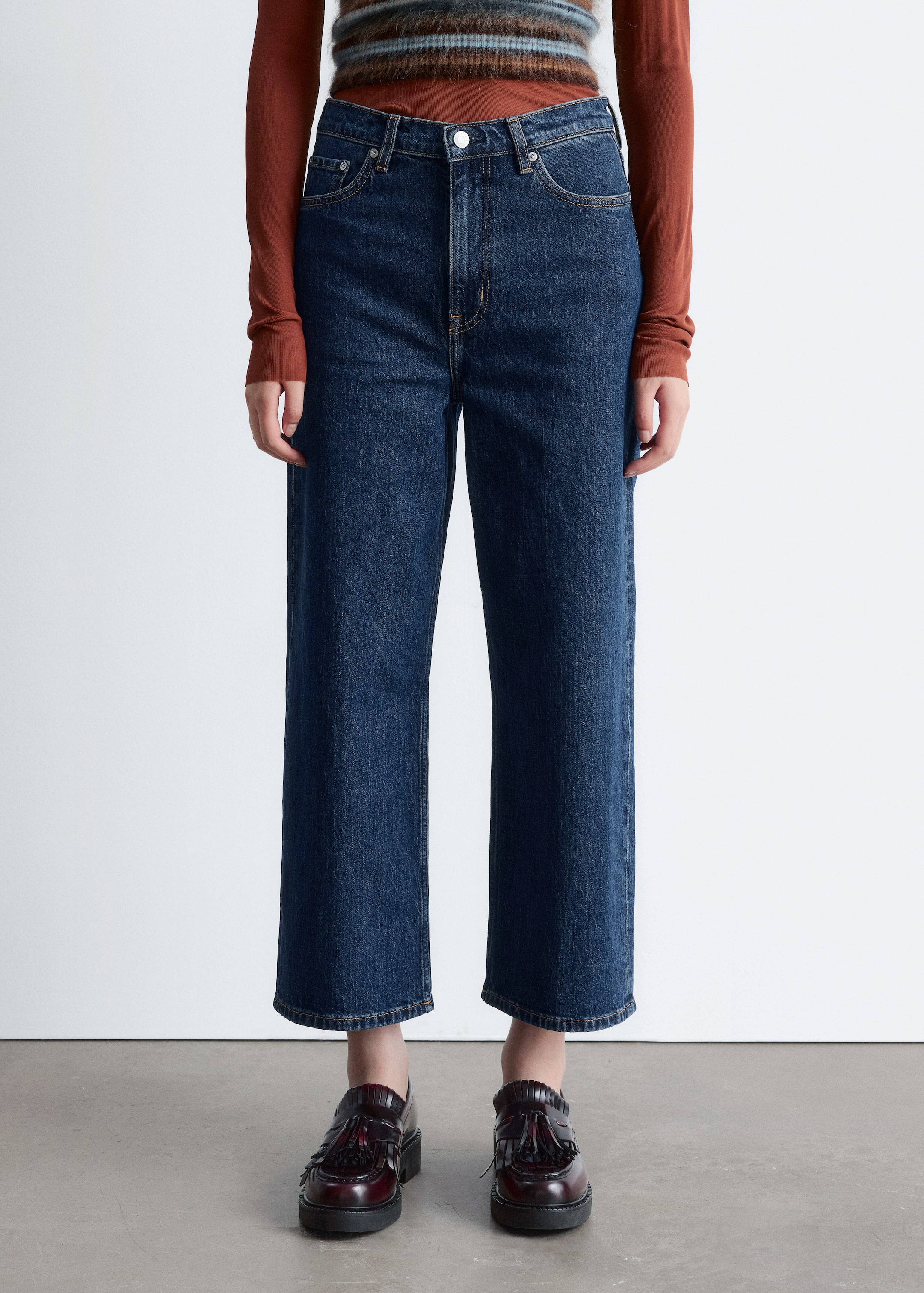 View larger image: Dark blue high-waisted jeans, featuring a 5-pocket design and cropped wide legs, are being worn by the model. The jeans are being styled with a long-sleeved top tucked in, highlighting the high waist, and with dark red loafers, emphasizing the ankle-length hem.