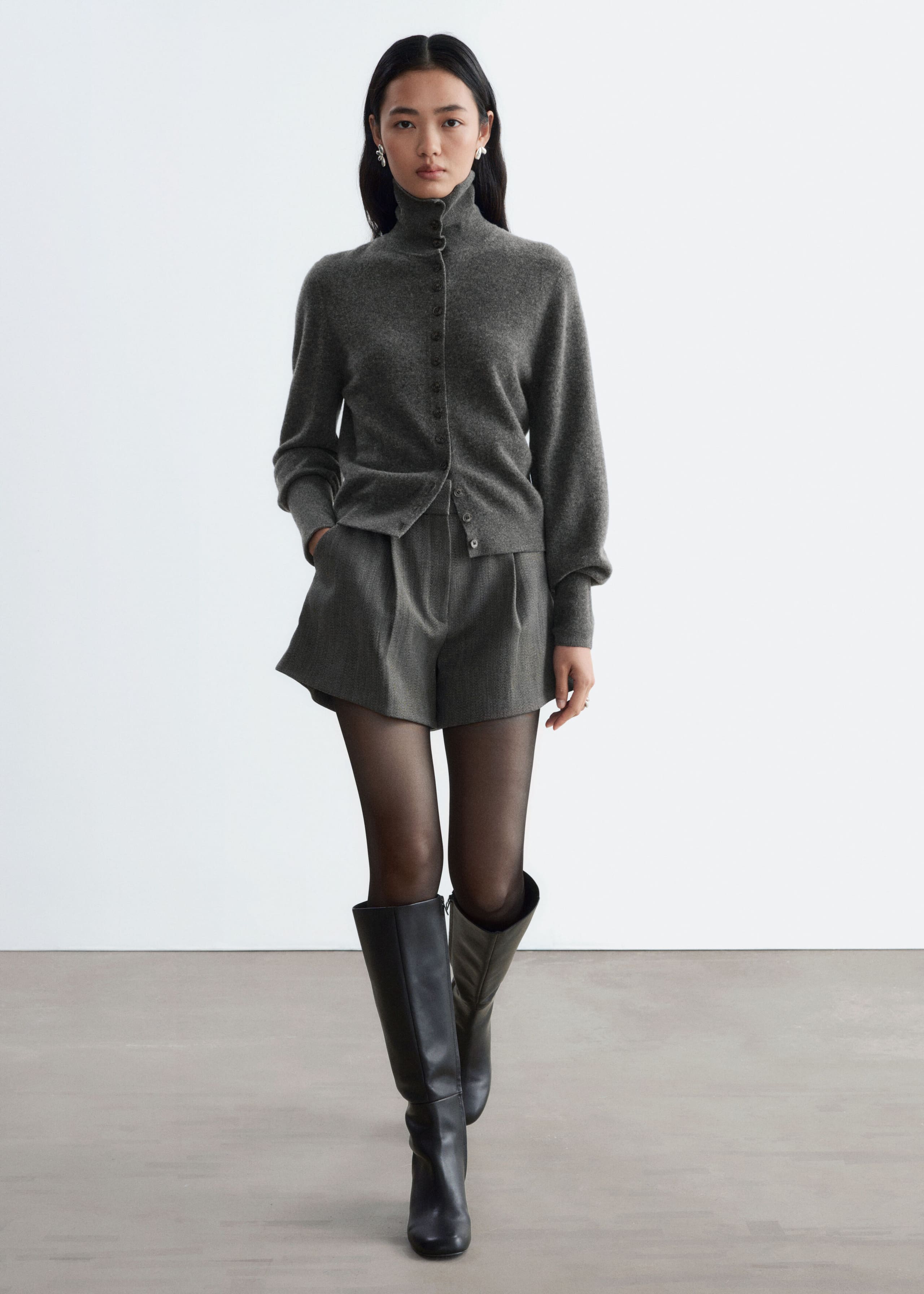 These gray, high-waisted wool-blend shorts feature front pleats and an A-line silhouette and are worn by the model, who stands with one hand resting in a side pocket. The shorts are styled with a matching gray high-neck cardigan, sheer black tights, and knee-high black leather boots.