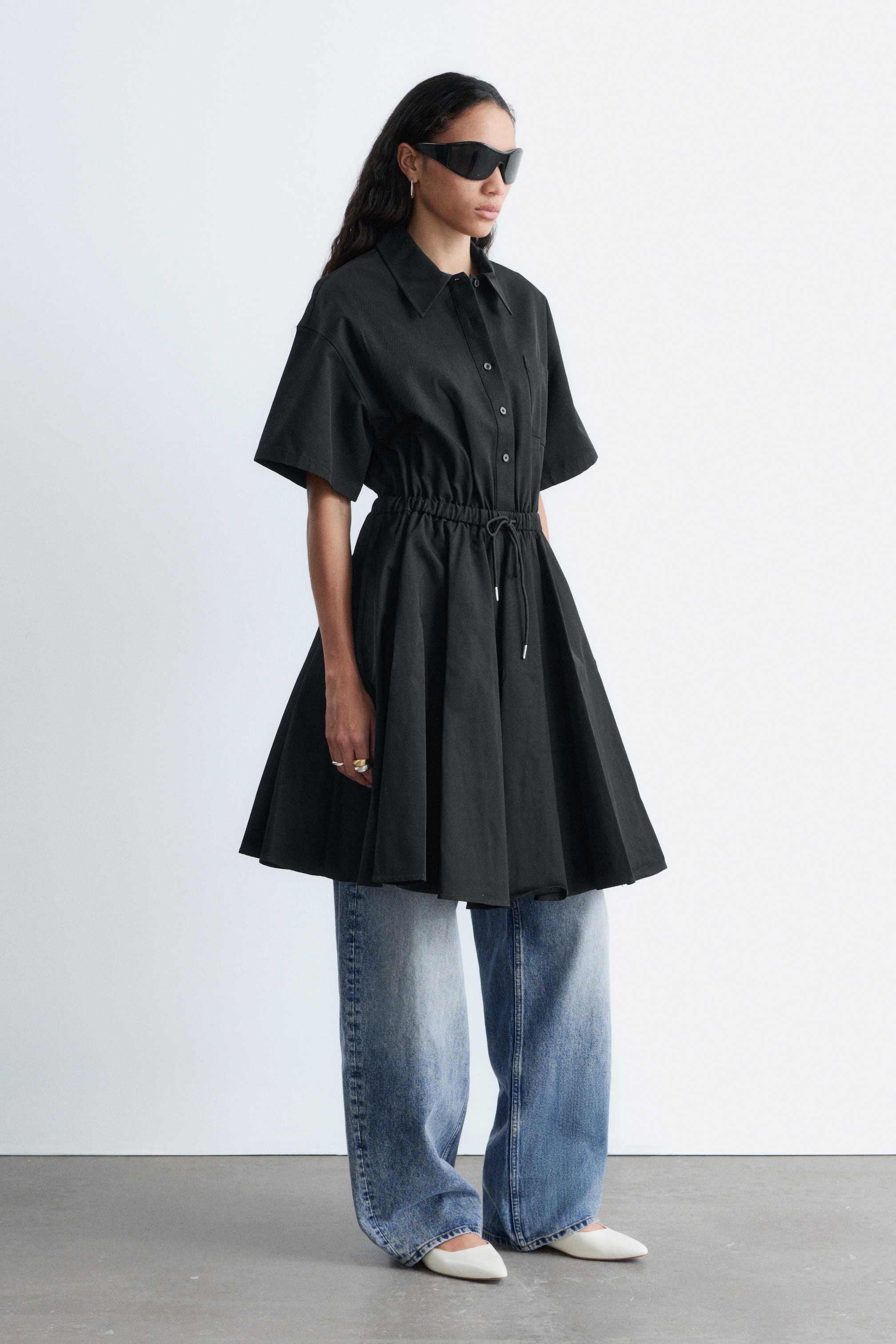 Ingrandisci l'immagine: Una tunica chemisier nera a maniche corte presenta un colletto, un'abbottonatura frontale e una tasca sul petto sinistro. Un cordoncino elasticizzato stringe la vita, creando una gonna svasata, ed è indossata a strati sopra jeans a gamba larga con lavaggio chiaro.