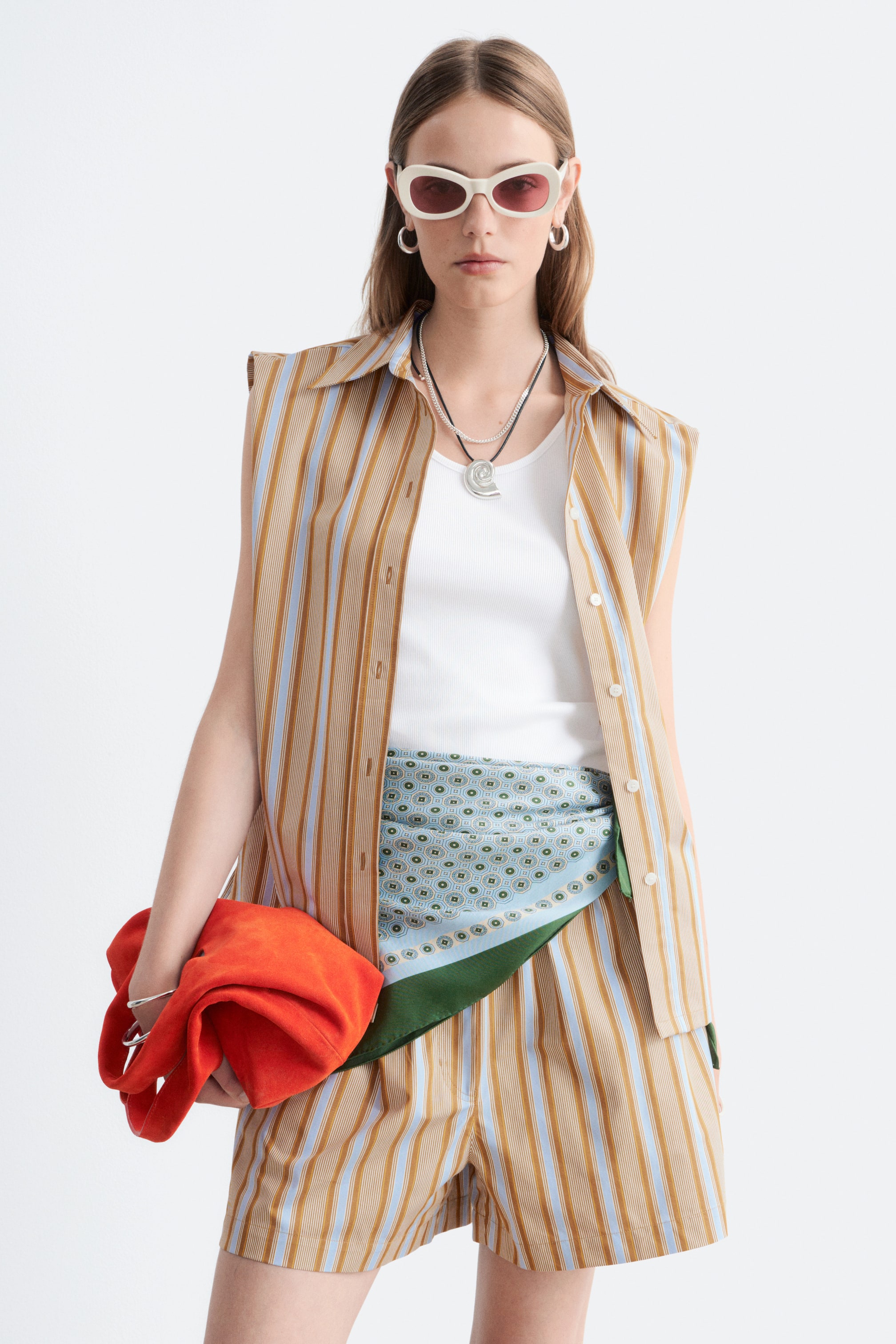 Agrandir l'image: Un modèle présente de face une chemise sans manches à rayures boutonnée et un short assorti. La chemise et le short arborent de fines rayures verticales en beige, bleu clair et marron ; la chemise est portée ouverte sur un débardeur côtelé blanc, et un foulard à motifs bleu clair est noué autour de la taille.
