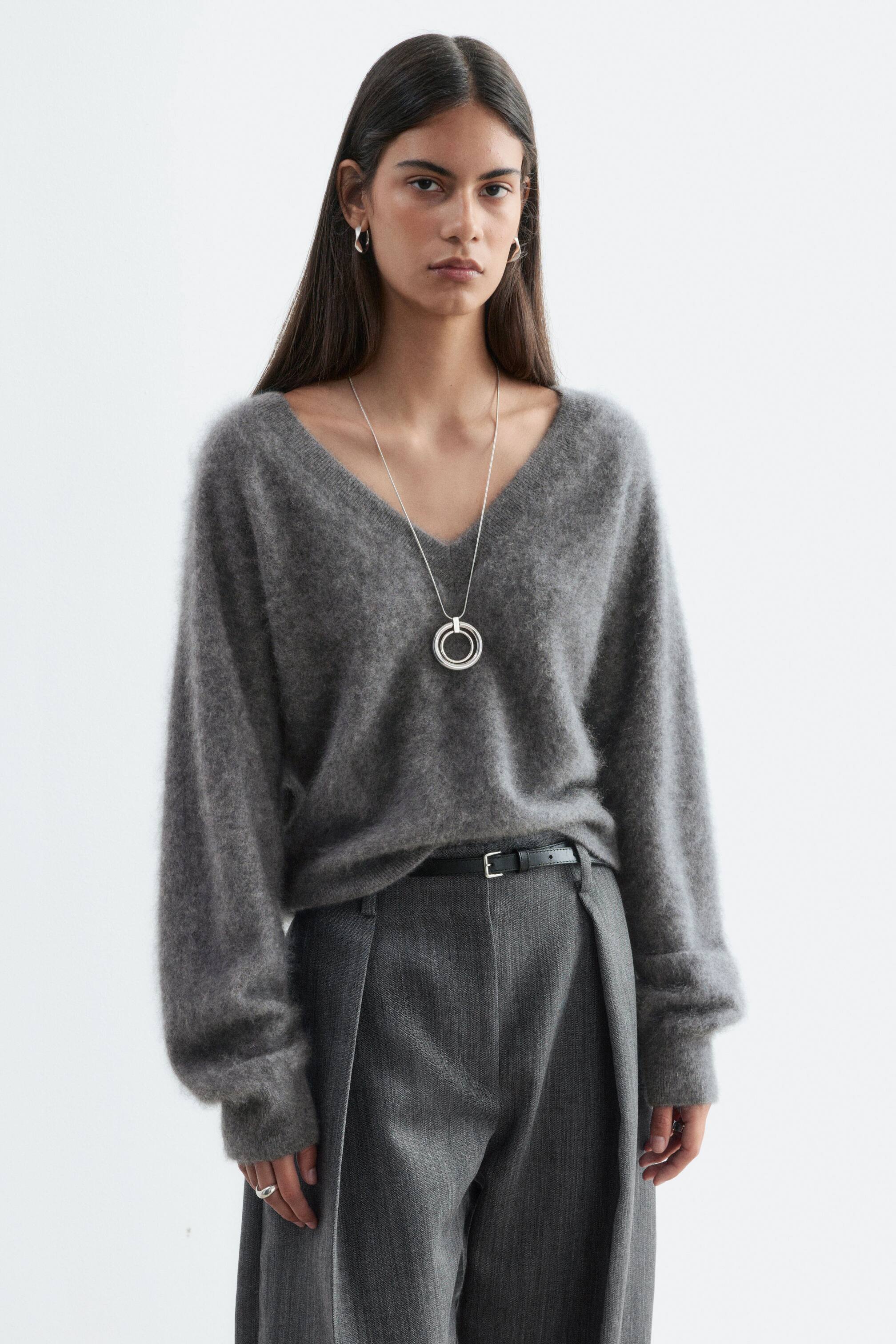 View larger image: A grey brushed cashmere V-neck jumper is worn by the model and tucked into high-waisted grey trousers. The long-sleeved textured knit is styled with a silver pendant necklace resting over the neckline.