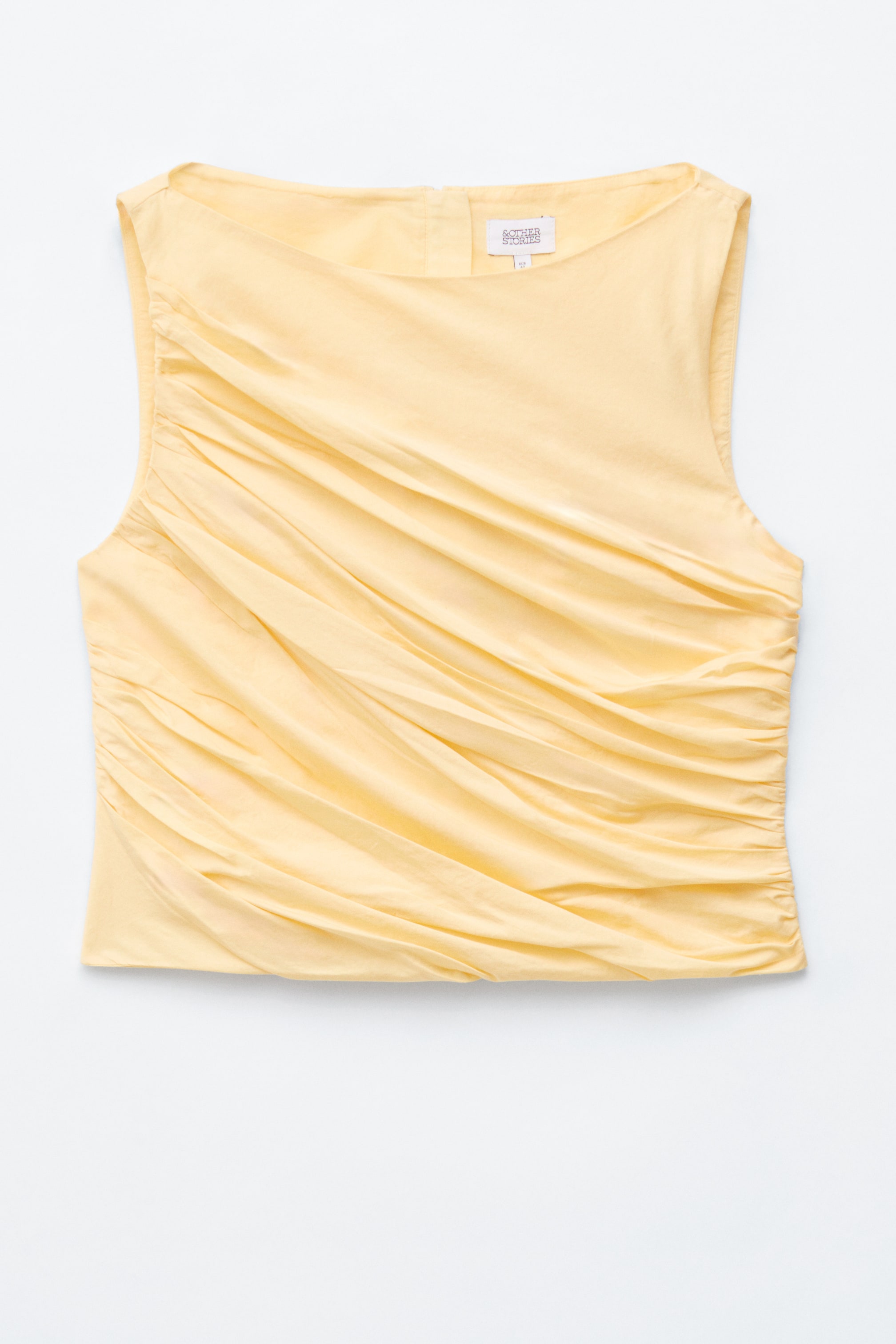 Nagyobb kép megtekintése: Egy halványsárga, ujjatlan felső látható az elülső oldaláról, amely csónaknyakú kivágással rendelkezik. Átlós ráncok gyűlnek össze a bal vállról a mellkasra, és a felső rövid hosszúságot mutat.