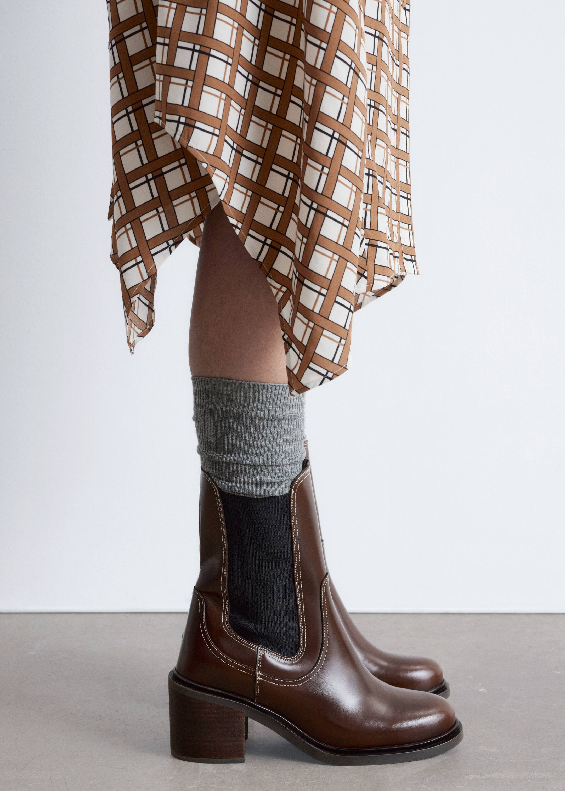 Agrandir l'image: Des bottines Chelsea en cuir marron sont présentées de profil, avec des panneaux latéraux élastiques noirs, des coutures contrastées blanches et un talon bloc empilé. Des chaussettes côtelées grises dépassent du haut des bottes.