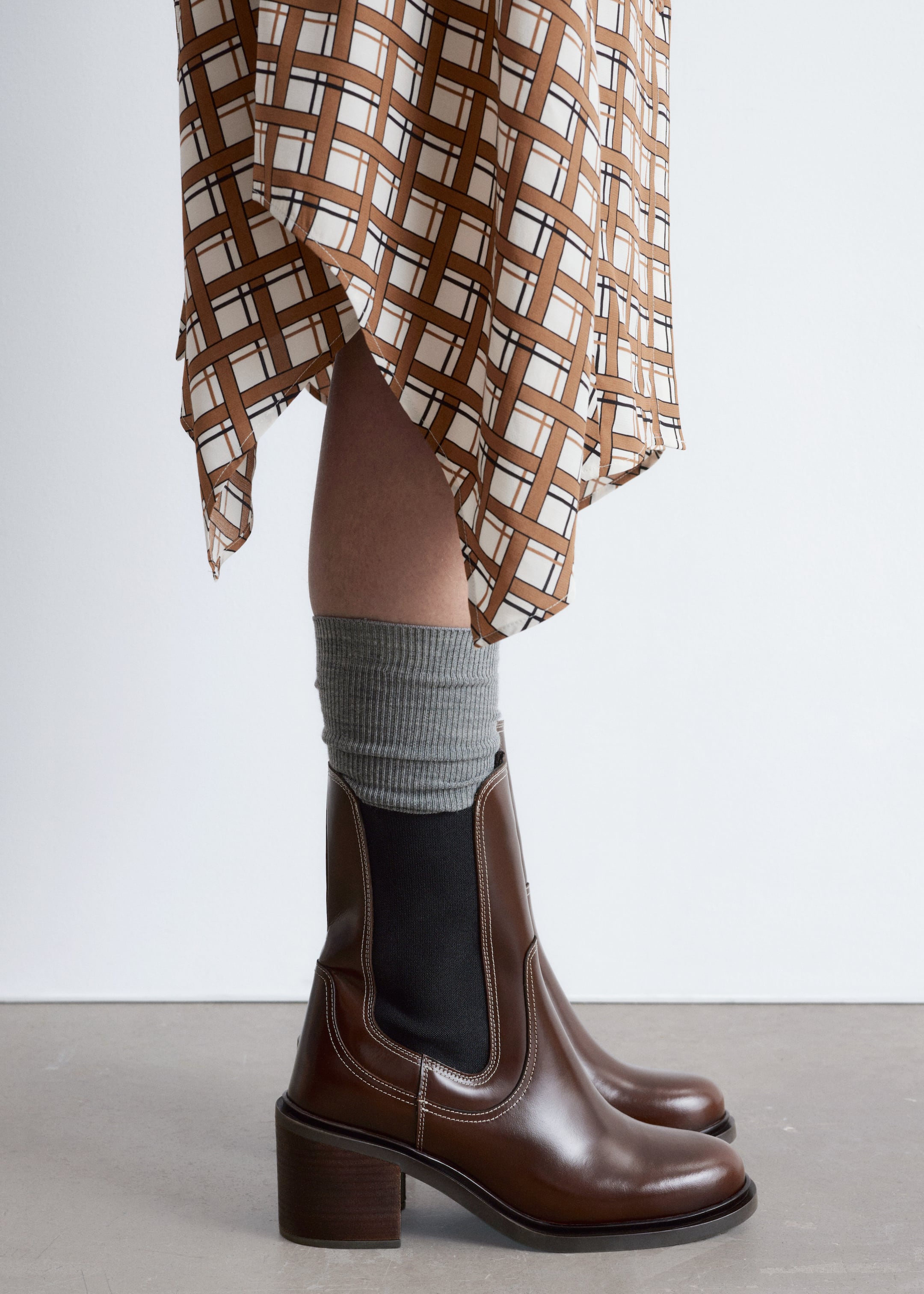 View larger image: Brown leather Chelsea boots are being presented in a side profile, featuring black elastic side panels, white contrast stitching, and a stacked block heel. Gray ribbed socks are peeking out from the top of the boots.
