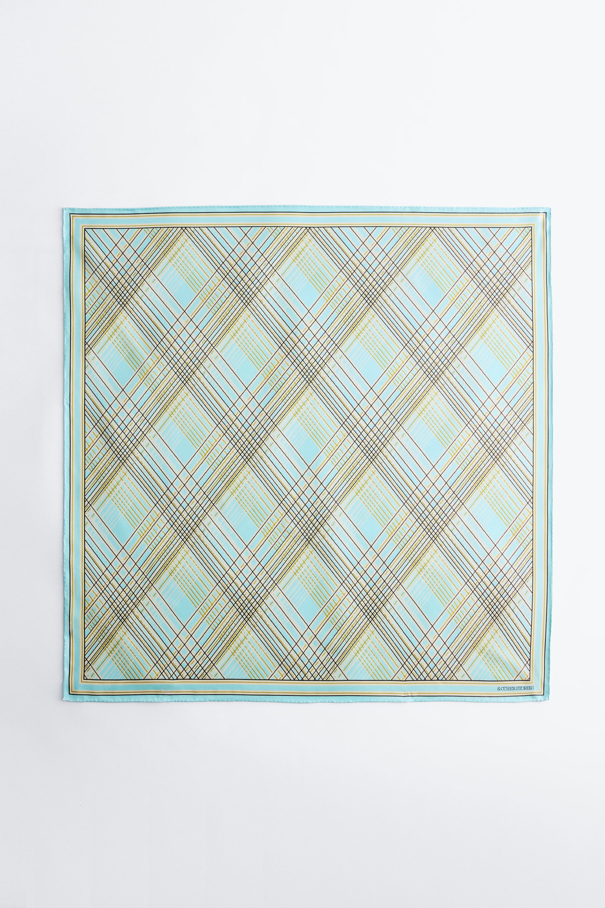 Größeres Bild ansehen: Bedrucktes Seidentuch. Das quadratische Seidentuch präsentiert sich mit einer hellblauen Basis und einem diagonalen Karomuster, das dünne braune, goldene und hellblaue Linien zeigt. Es ist mit einem passenden gestreiften Rand versehen, und ein kleines „& OTHER STORIES“-Logo ist unten rechts sichtbar.
