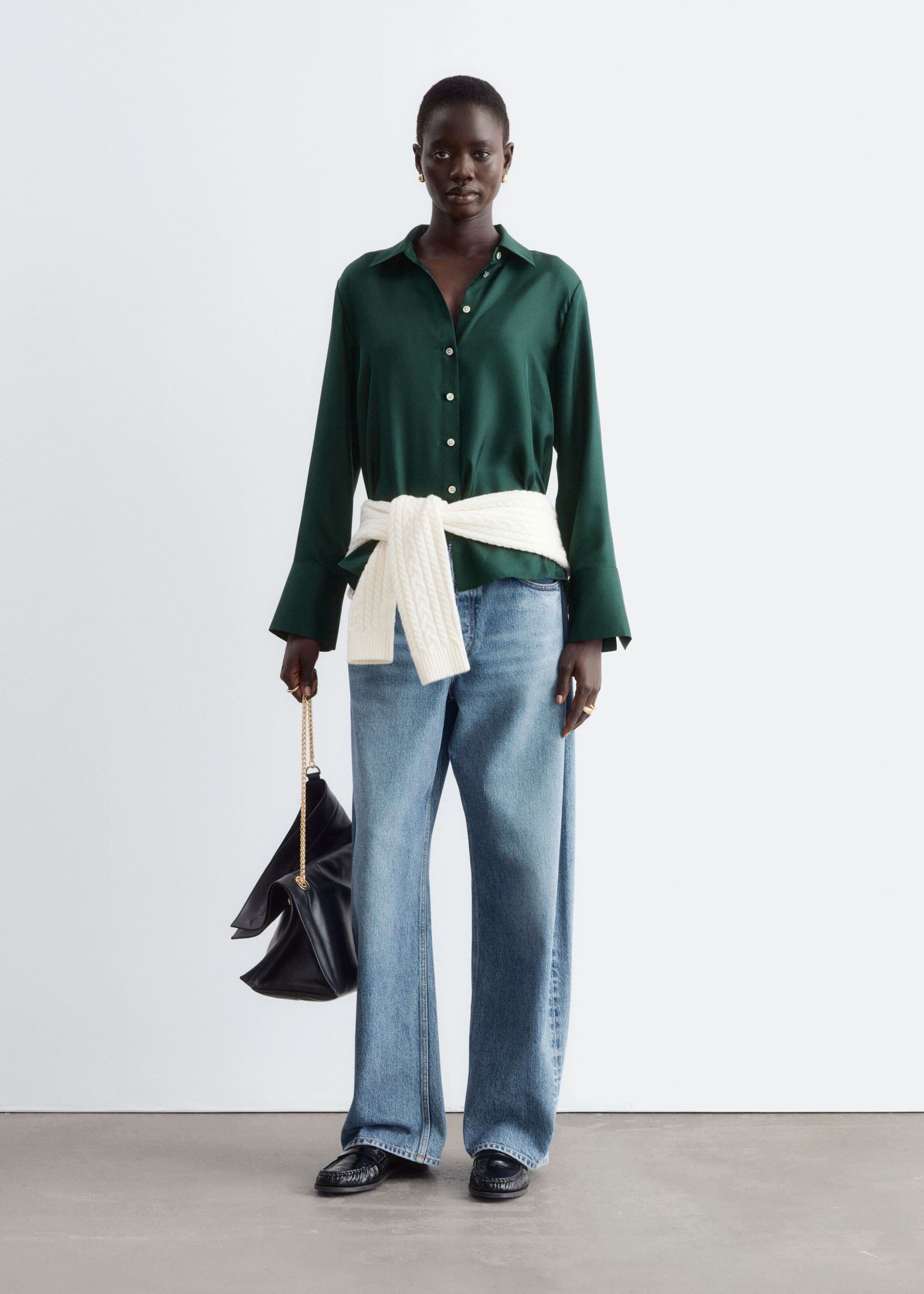A dark green silk shirt with a classic collar, button-down front, and wide cuffs is worn by the model. The shirt is styled with light-wash, wide-leg jeans, a white cable-knit jumper tied around the waist, and a black handbag held in one hand.