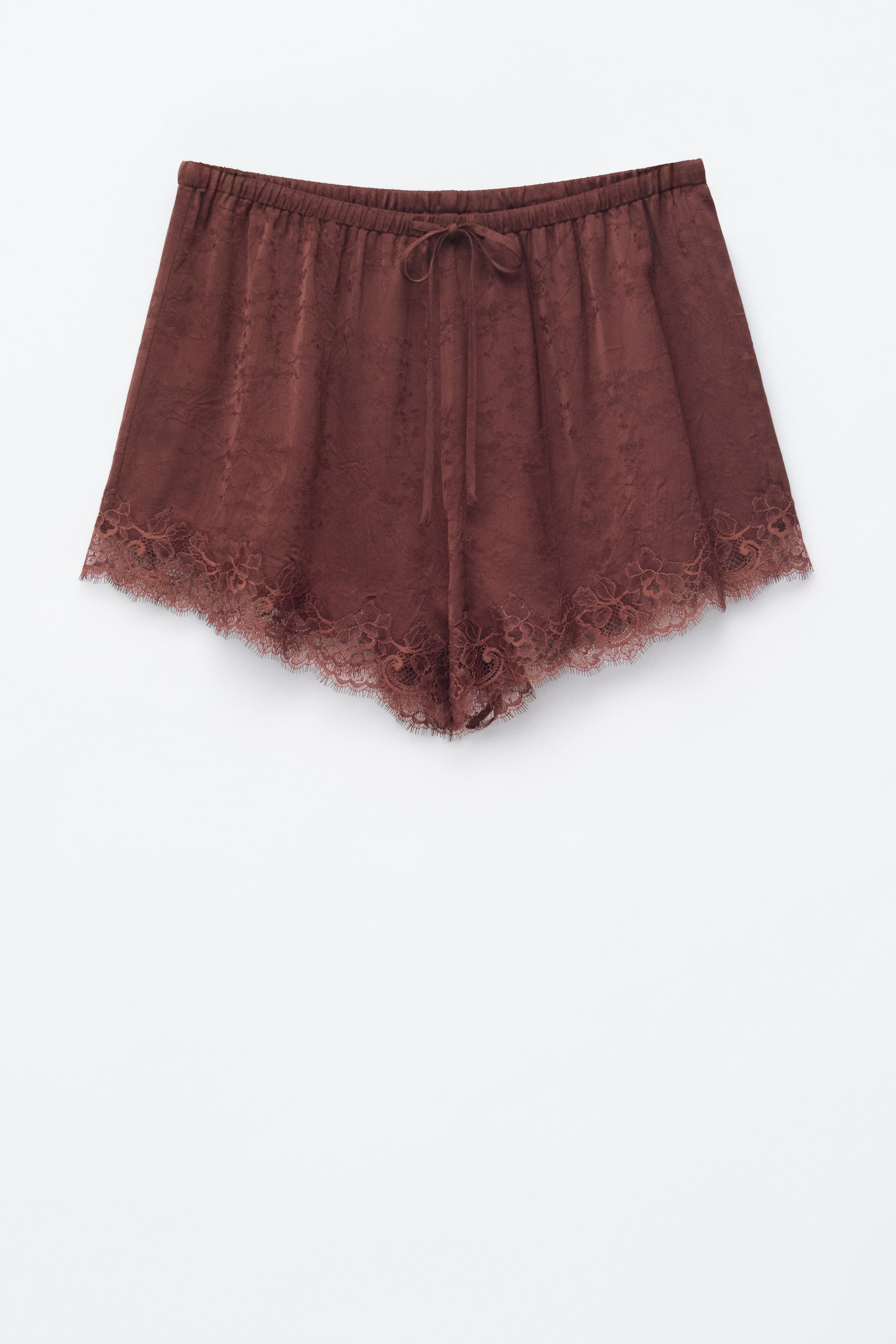 Visualizza immagine più grande: I pantaloncini marrone scuro sono distesi in piano e rivolti in avanti, caratterizzati da una vita elasticizzata con un cordoncino annodato a fiocco. Un sottile motivo jacquard è visibile sul loro tessuto setoso, e le aperture delle gambe sono rifinite con un pizzo floreale smerlato.
