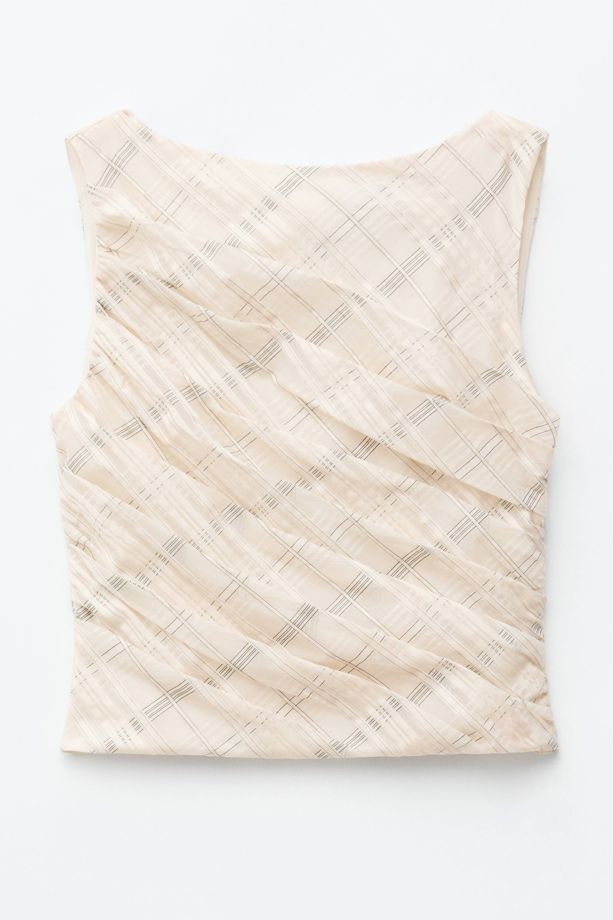 Agrandir l'image: Un haut sans manches est présenté de face, arborant une couleur beige clair avec un motif à carreaux subtil. Des plis artisanaux en diagonale créent texture et profondeur sur l'ensemble du vêtement, tandis qu'un large col bateau et une courte longueur sont visibles.