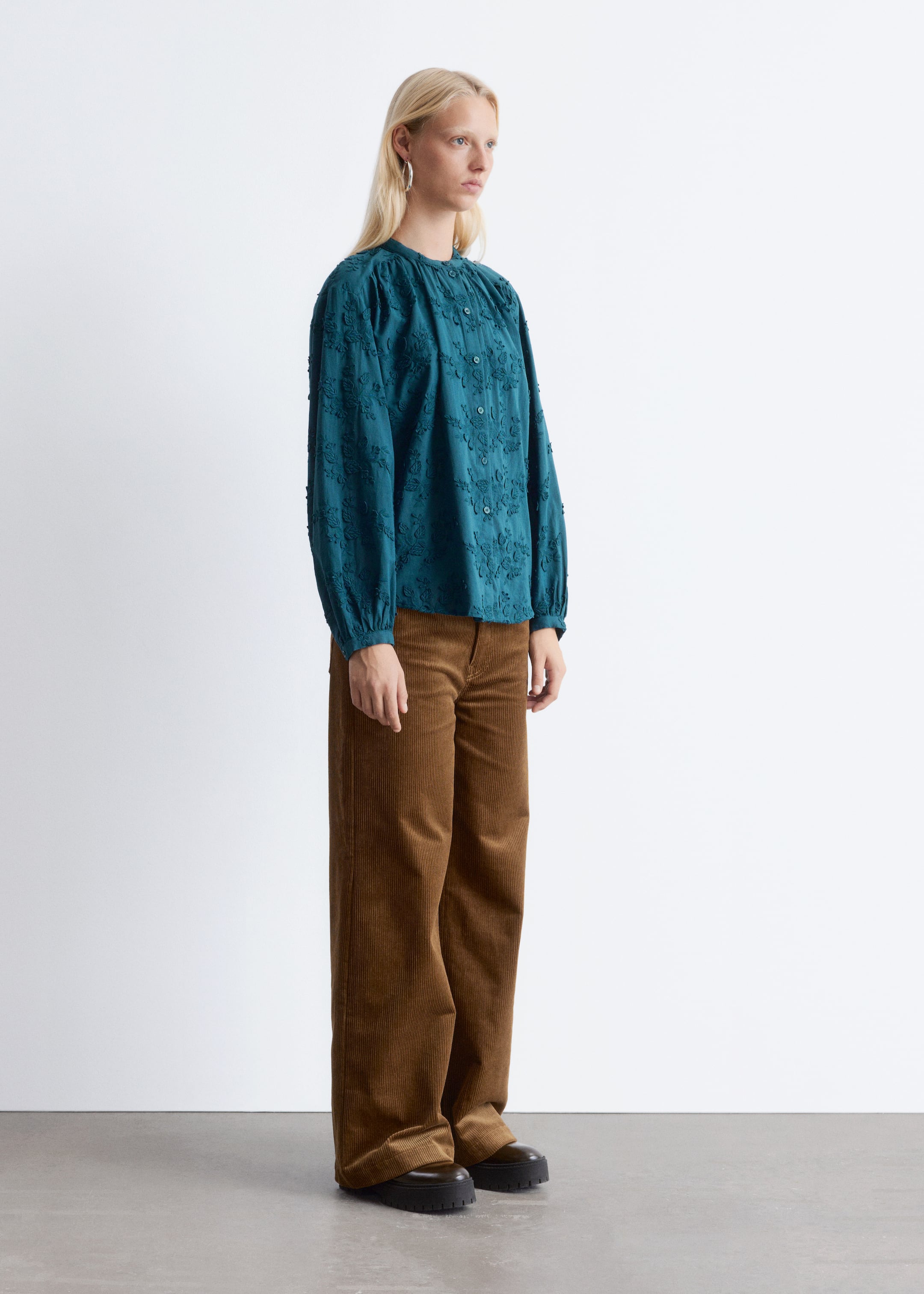 Visualizza immagine più grande: Una blusa verde petrolio scuro, caratterizzata da maniche lunghe arricciate e un motivo floreale ricamato, viene presentata in vista laterale. È abbinata a pantaloni a gamba larga in velluto a coste marrone e scarpe scure con suola spessa.