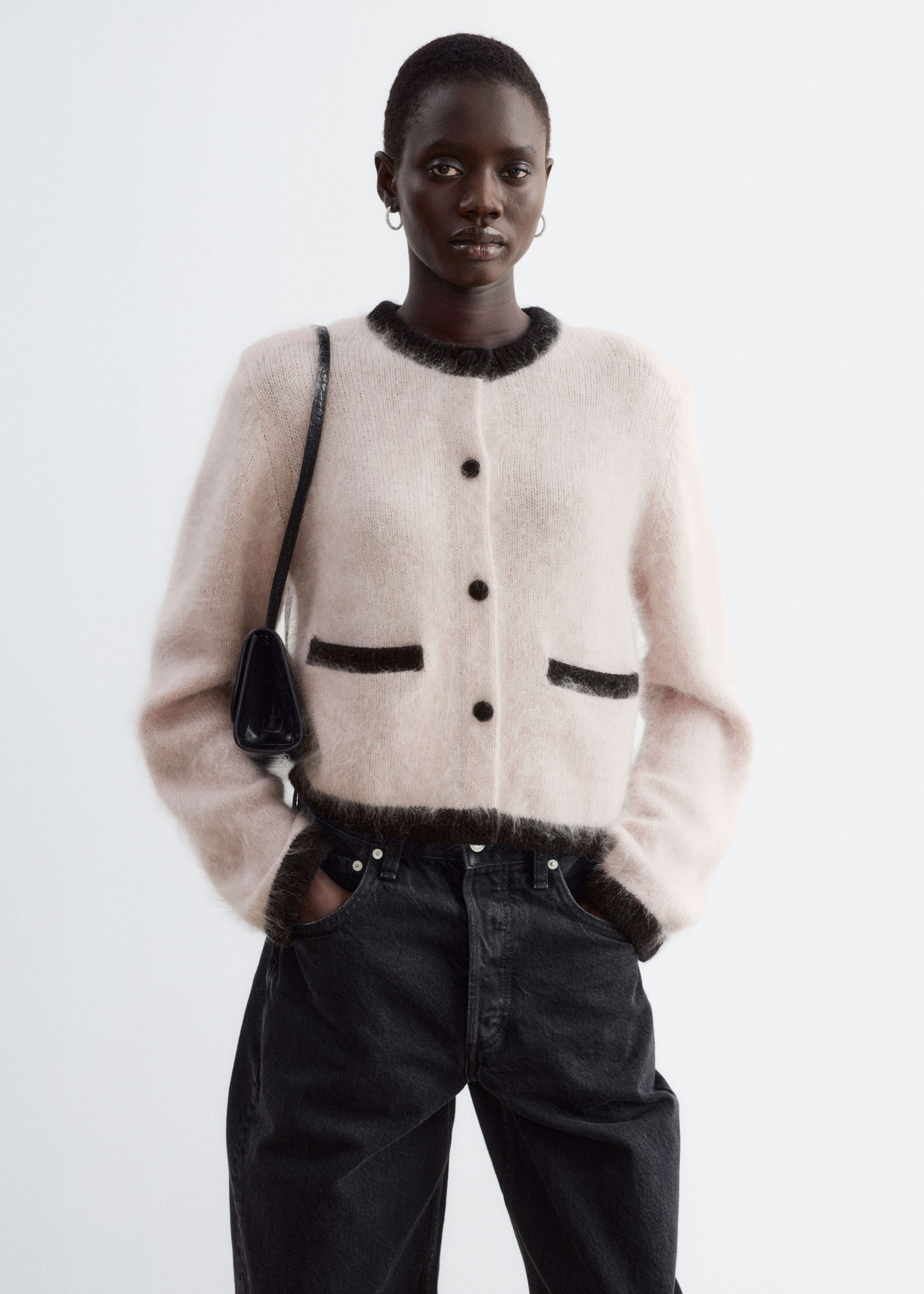 A fuzzy, light pink cardigan with structured shoulders and contrasting dark ribbed trim is being worn by the model. The cardigan features dark front buttons and mock pocket details, and is styled with dark-wash jeans and a black shoulder bag.