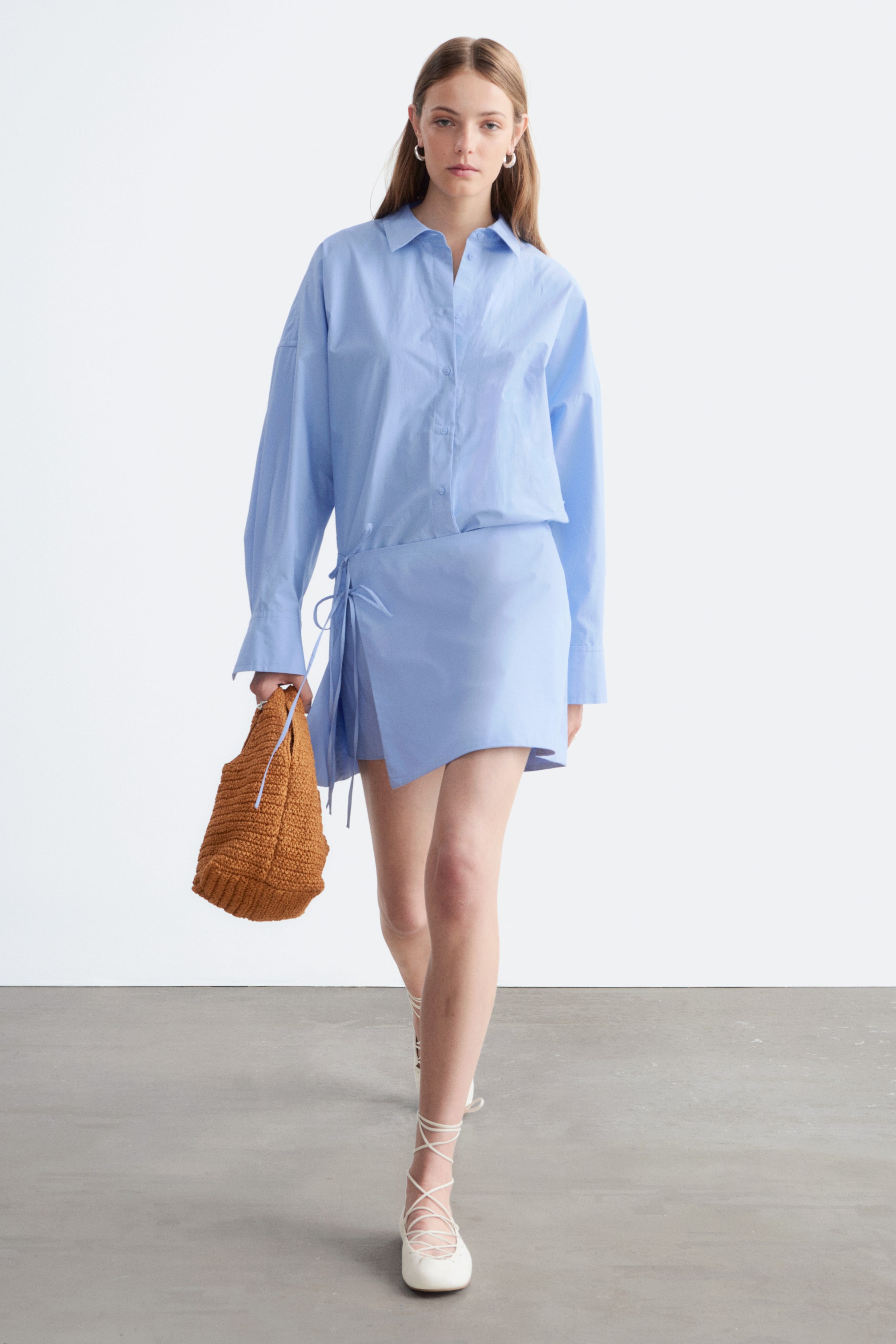 Agrandir l'image: Une chemise bleu clair à col, avec des manches longues et des poignets boutonnés, est portée de manière ample et partiellement rentrée dans une mini-jupe portefeuille assortie bleu clair. La jupe présente des liens visibles sur son côté gauche et un ourlet asymétrique.