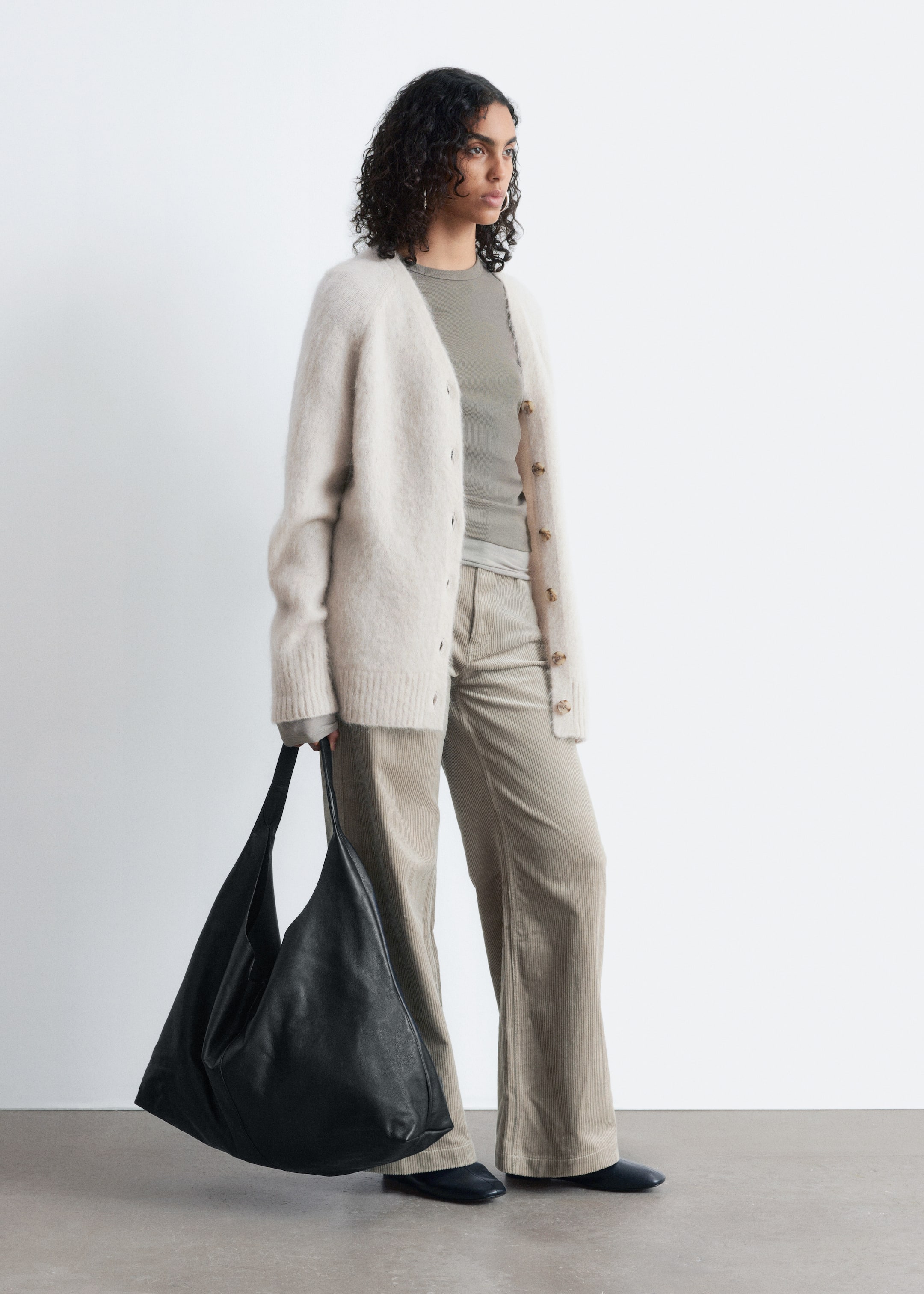 Visualizza immagine più grande: Il Cardigan Lungo in Misto Mohair, beige chiaro con una texture pelosa e bottoni marroni, è indossato aperto dalla modella. È stratificato sopra una maglietta a maniche lunghe verde chiaro, che arriva sotto i fianchi.
