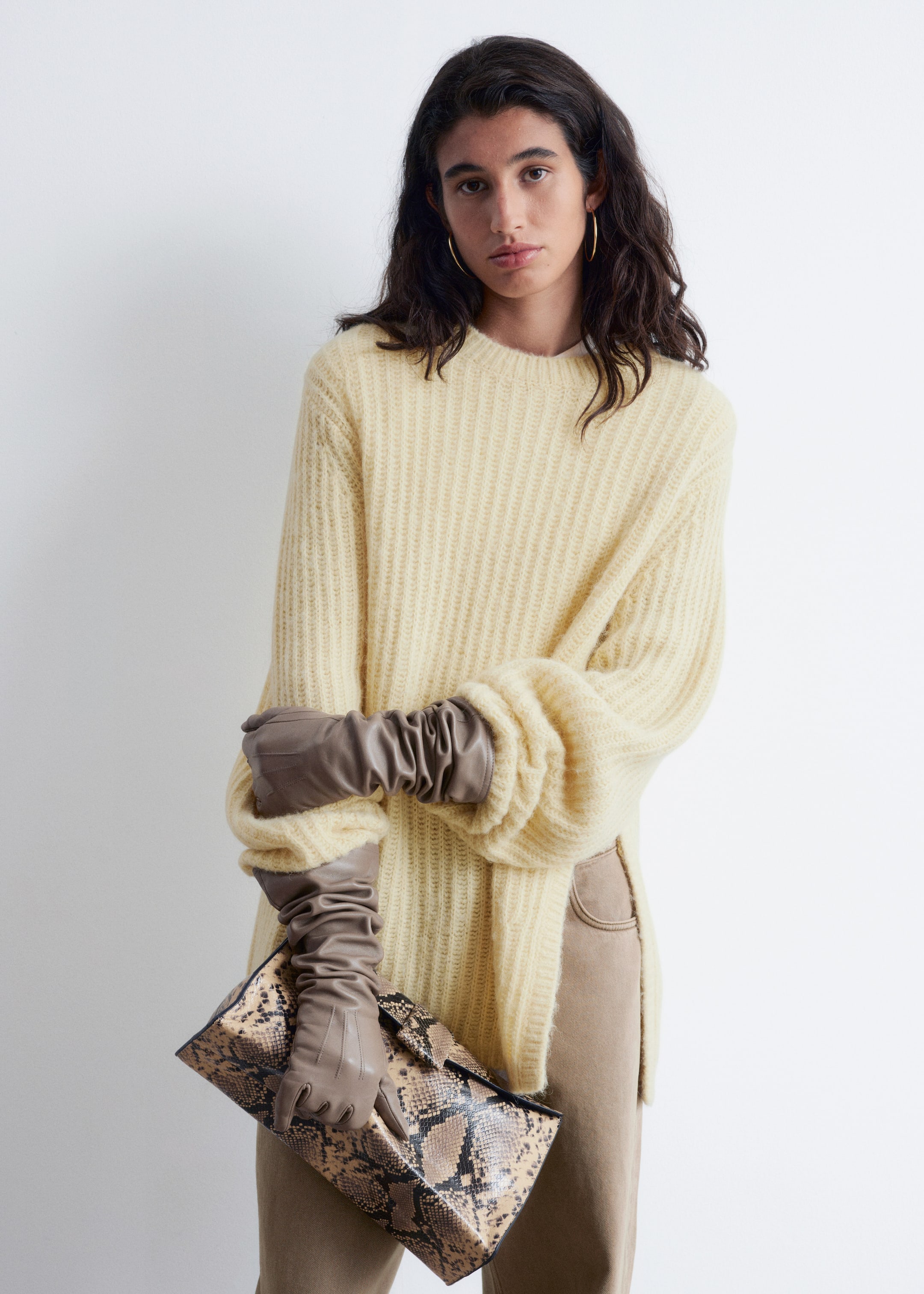 Visualizza immagine più grande: Un maglione a coste di colore giallo pallido con scollo rotondo viene presentato, caratterizzato da maniche lunghe voluminose che si raccolgono su guanti arricciati lunghi, e spacchi laterali sull'orlo che rivelano pantaloni beige sottostanti.