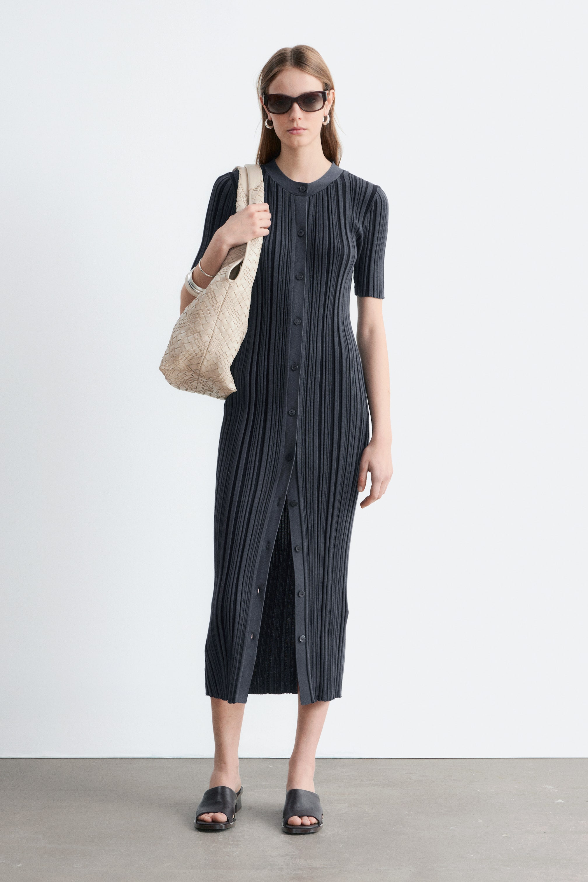 Visualizza immagine più grande: Un abito midi a coste grigio scuro, caratterizzato da una silhouette aderente, scollo rotondo, maniche corte e chiusura frontale con bottoni, con alcuni bottoni inferiori slacciati per creare uno spacco anteriore, è indossato dalla modella. La modella porta anche una borsa intrecciata di colore chiaro e indossa dei sabot scuri.