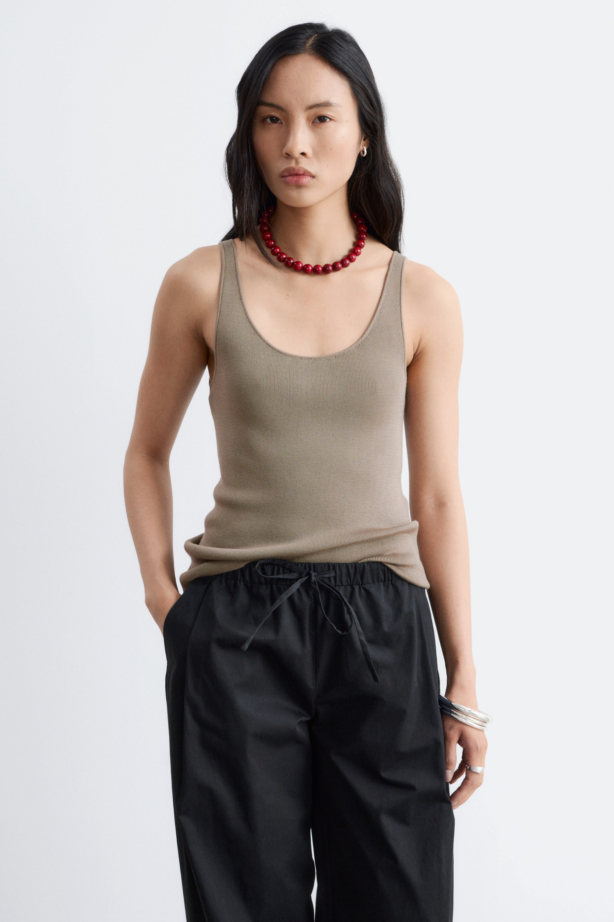 Vezi imaginea mai mare: Un maiou ribbed de culoare taupe, cu un decolteu adânc în formă de scoică și o țesătură fină, este purtat de model. Este stilizat neîndesat peste pantaloni negri, cu un colier din mărgele roșii așezat pe partea din față.