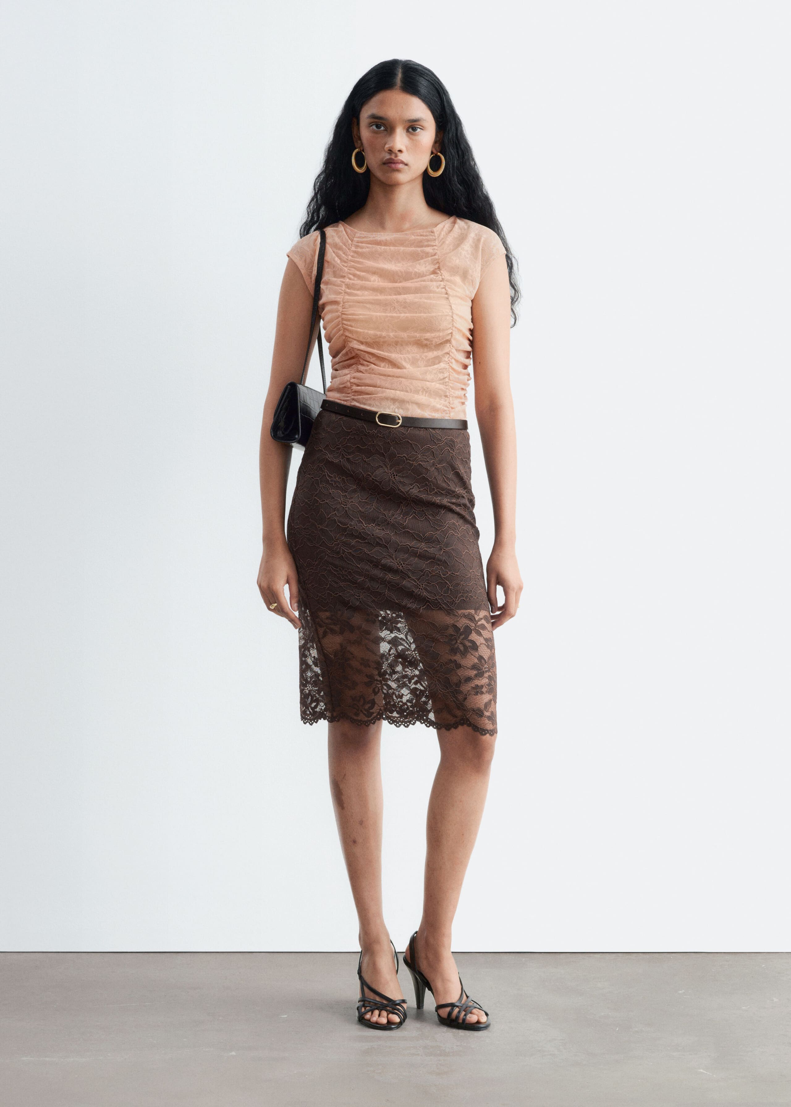 A peach-coloured lace top featuring ruched detailing through the body, cap sleeves, and a boat neckline is being worn by the model. The top is tucked into a brown lace skirt styled with a thin belt.