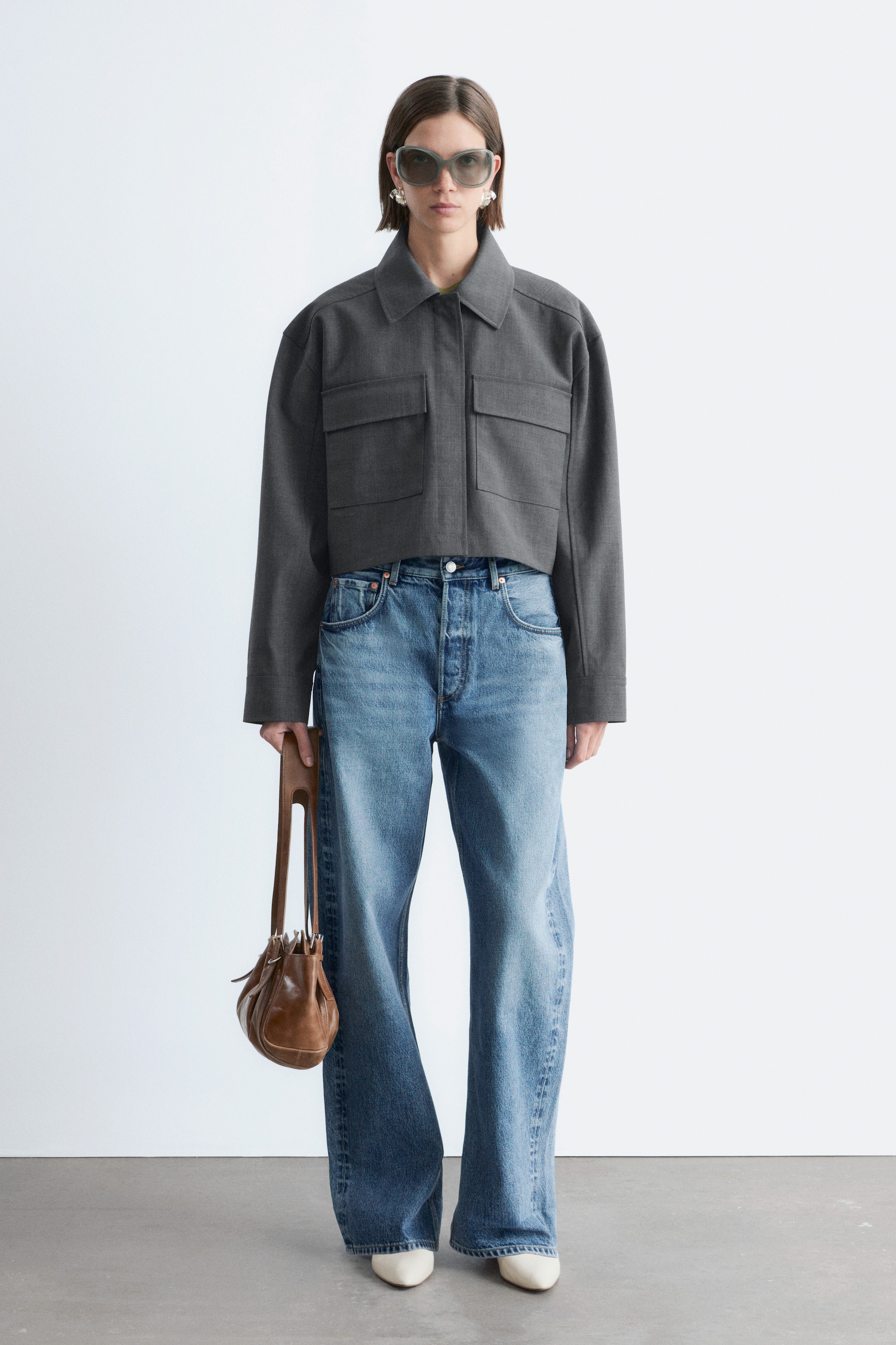 Se større billede: En mørkegrå kort jakke viser en spredt krave, lange ærmer, to brystlommer med klap og en boxy silhuet. Den er placeret med fronten fremad.