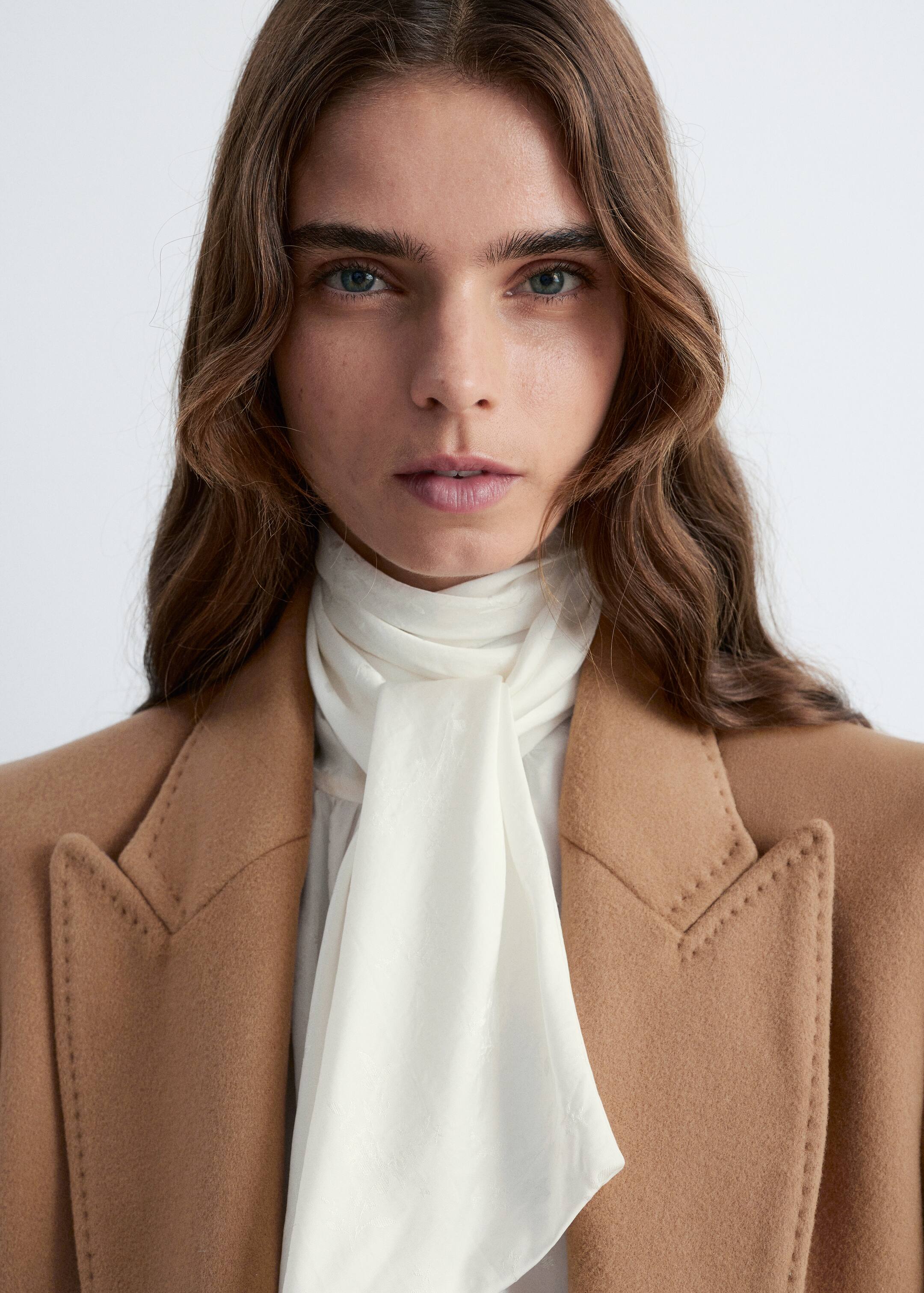 View larger image: A camel-colored coat with prominent notched lapels and decorative topstitching is being worn over a white blouse featuring a long, flowing pussy-bow tie, its lightweight fabric draping down the center.