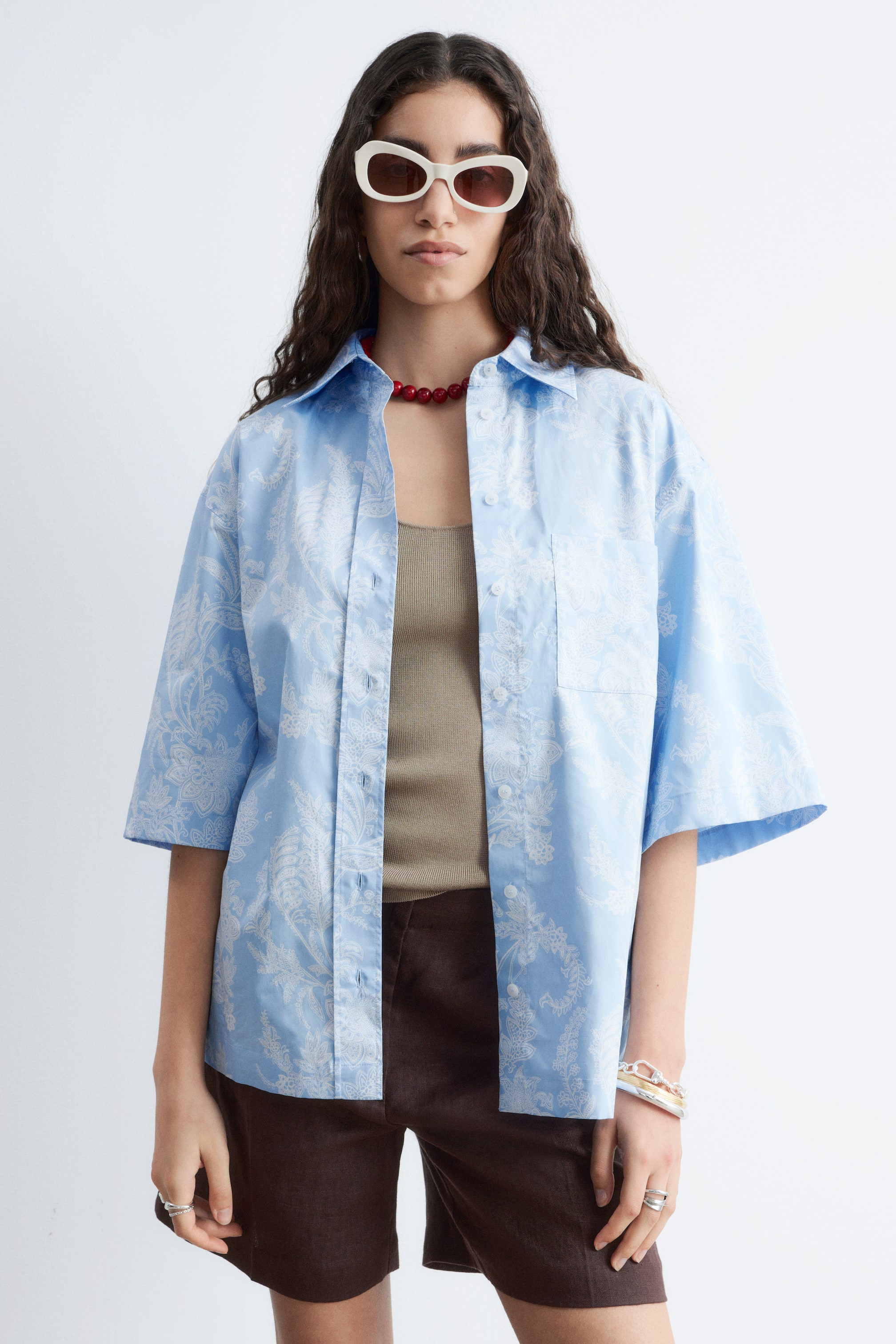 View larger image: A light blue short-sleeved shirt with a white paisley-like pattern featuring a classic collar, a button-up front with white buttons, and a single chest pocket on the left side. It presents a loose fit, worn open over a brown vest top.