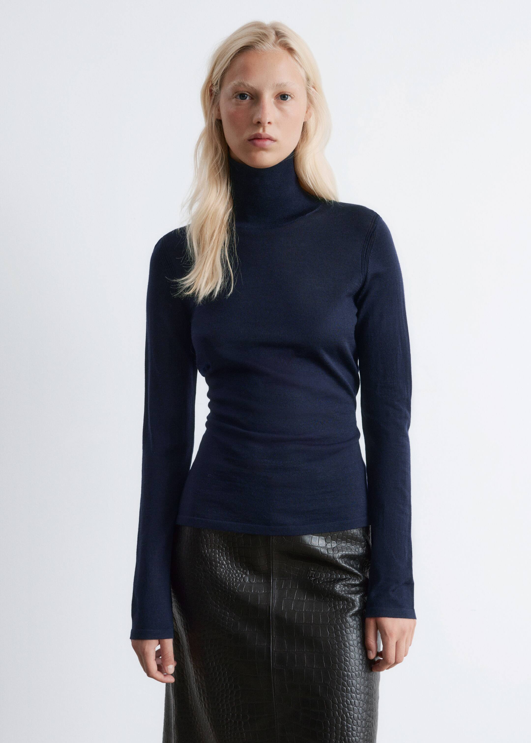 View larger image: A dark blue turtleneck top is being worn, facing forward, showcasing its slim fit. It is featuring a high, folded turtleneck, long fitted sleeves, and visible seam detailing along the shoulders. The top is extending to the upper hips, being styled over a black crocodile-embossed skirt.