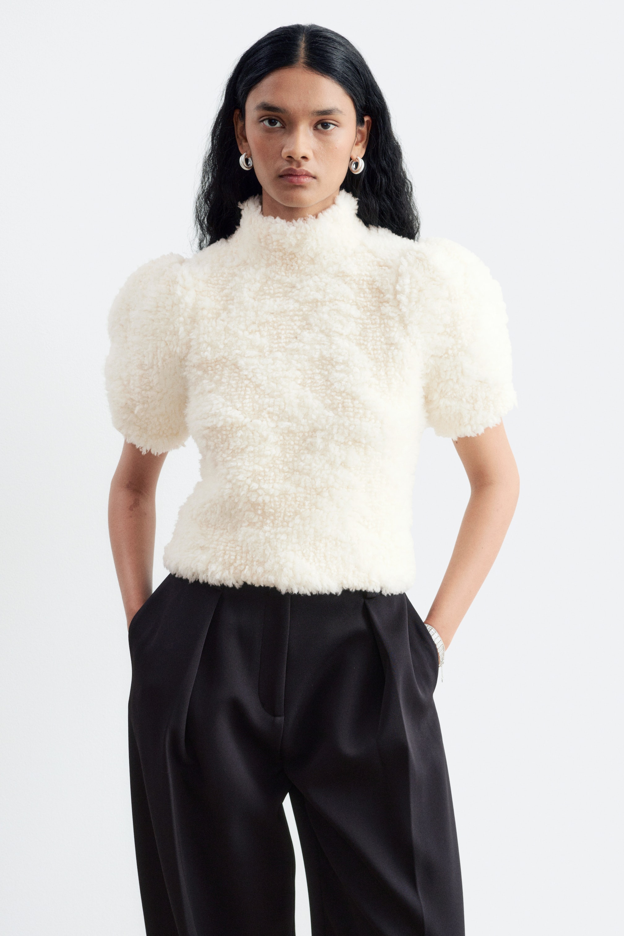 Visualizza l'immagine ingrandita: La modella indossa un top in maglia color crema caratterizzato da una superficie altamente strutturata e ad anelli, con collo alto finto e maniche corte a palloncino. Il top è abbinato a pantaloni neri plissettati a vita alta.