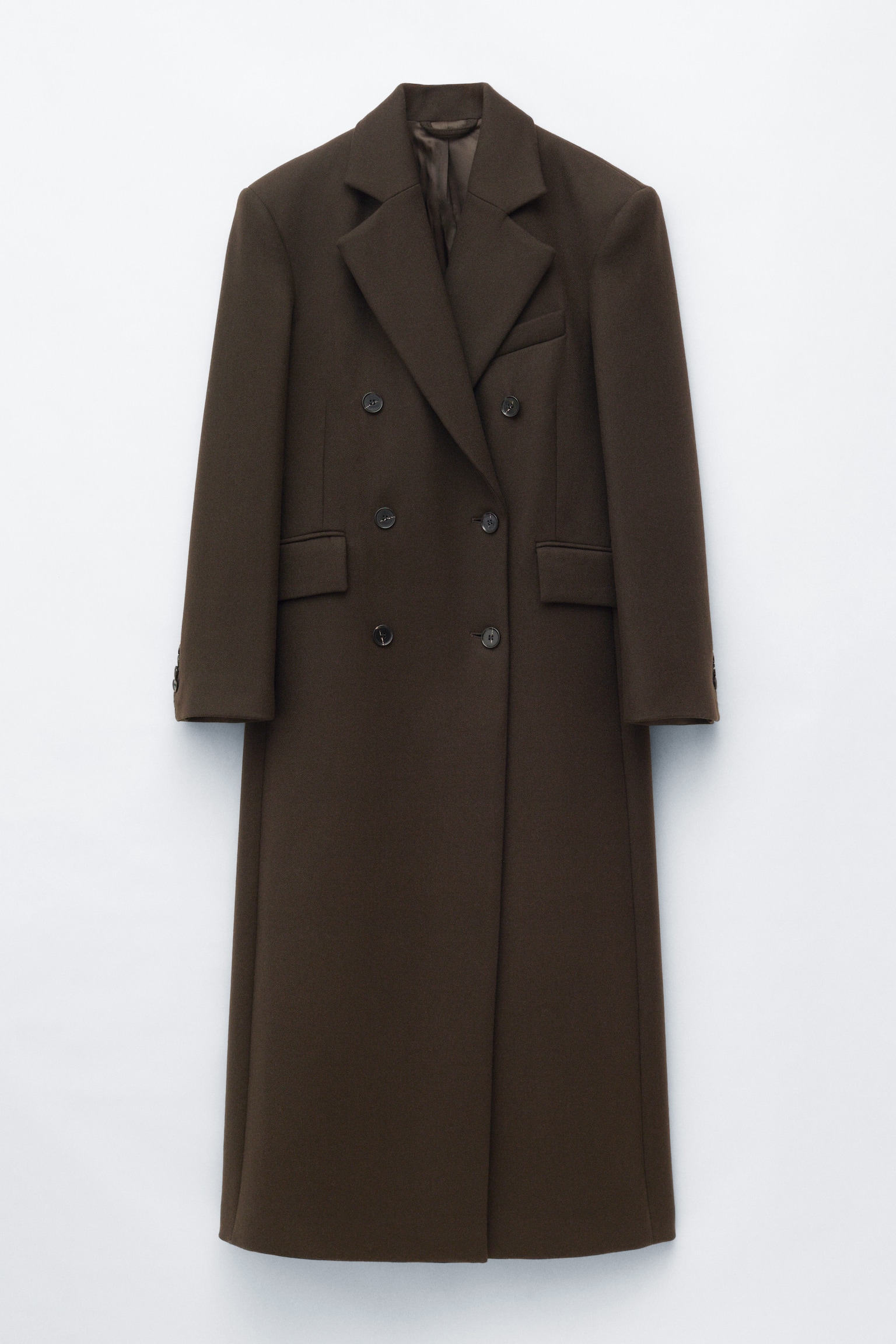 Un long manteau croisé marron foncé est suspendu de face, mettant en valeur une silhouette droite avec des épaules structurées et de larges revers crantés. Le manteau présente une fermeture frontale à six boutons, des poches latérales à rabat et une seule poche passepoilée sur la poitrine. - 1