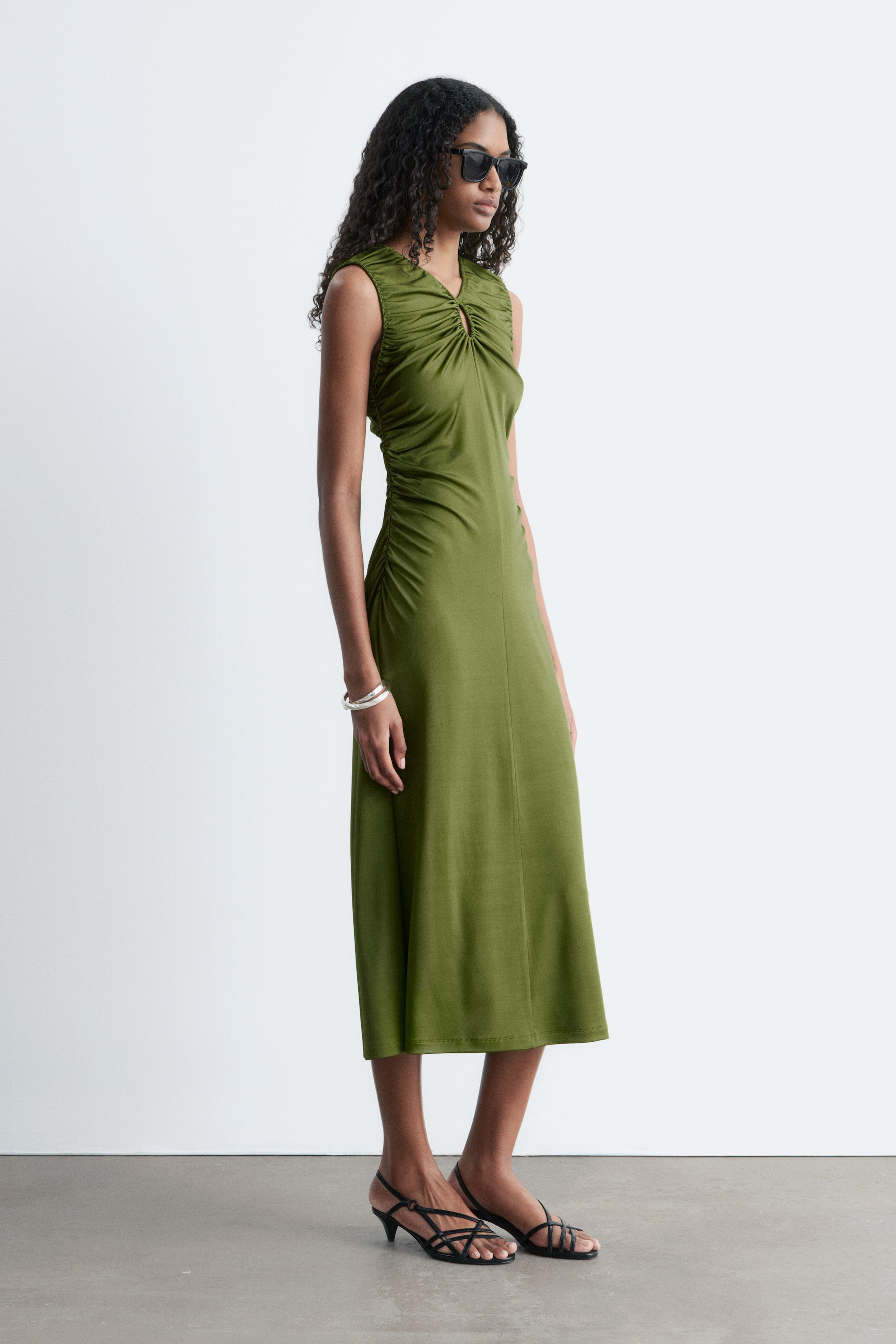 View larger image: An olive green sleeveless midi dress featuring a V-neckline with a central keyhole and gathered detailing at the bust. The dress showcases ruched sides and a flowing midi skirt, presented in a side profile.