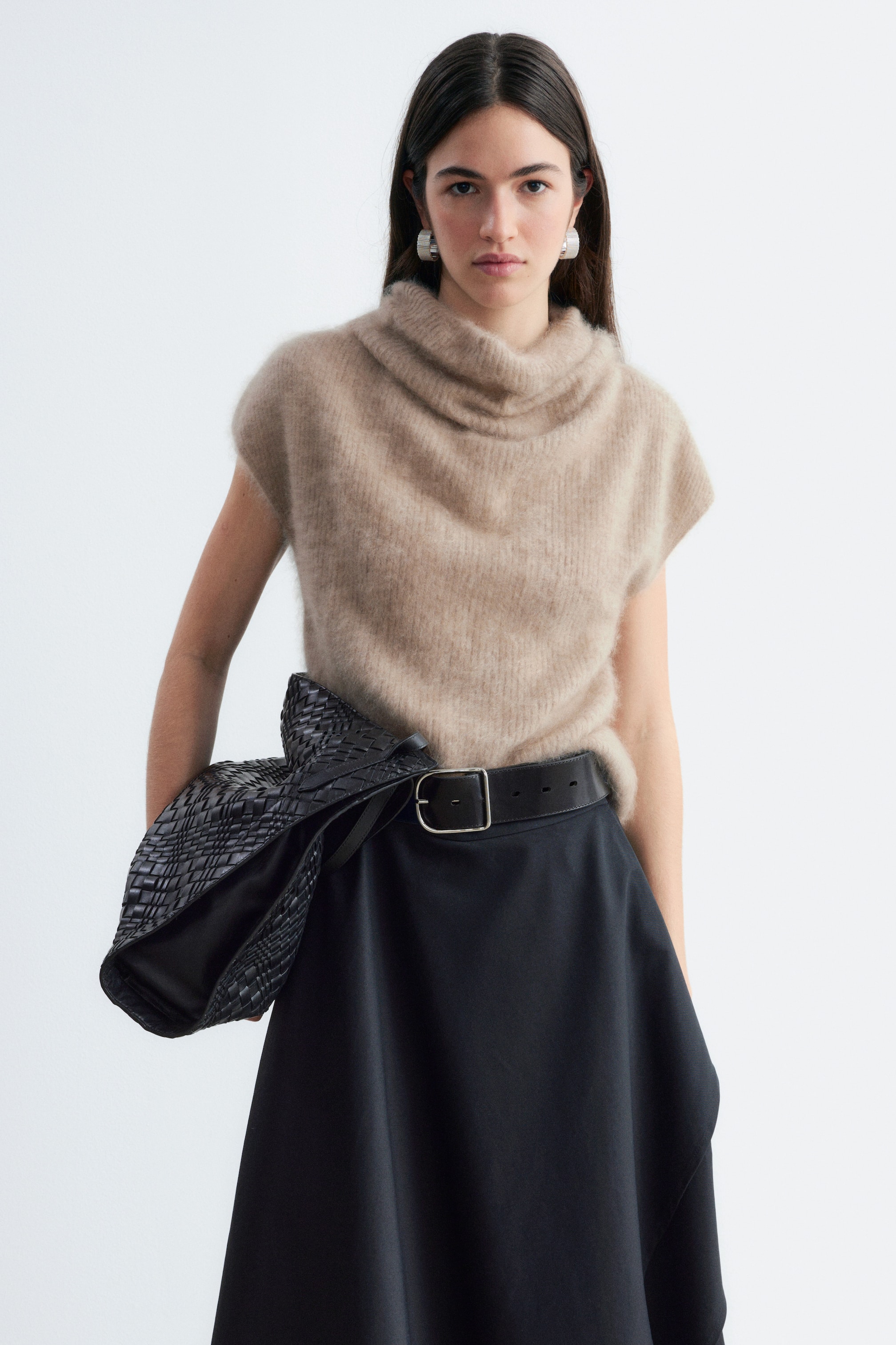 Se større billede: Cashmere Rib Mock Neck-toppen i en beige, fnugget strik bæres af modellen og har en draperet mock-neck-halsudskæring samt korte cap-ærmer. Toppen er stukket ned i et sort nederdel, der er holdt på plads af et bredt sort bælte, mens en sort flettet taske bæres under armen.