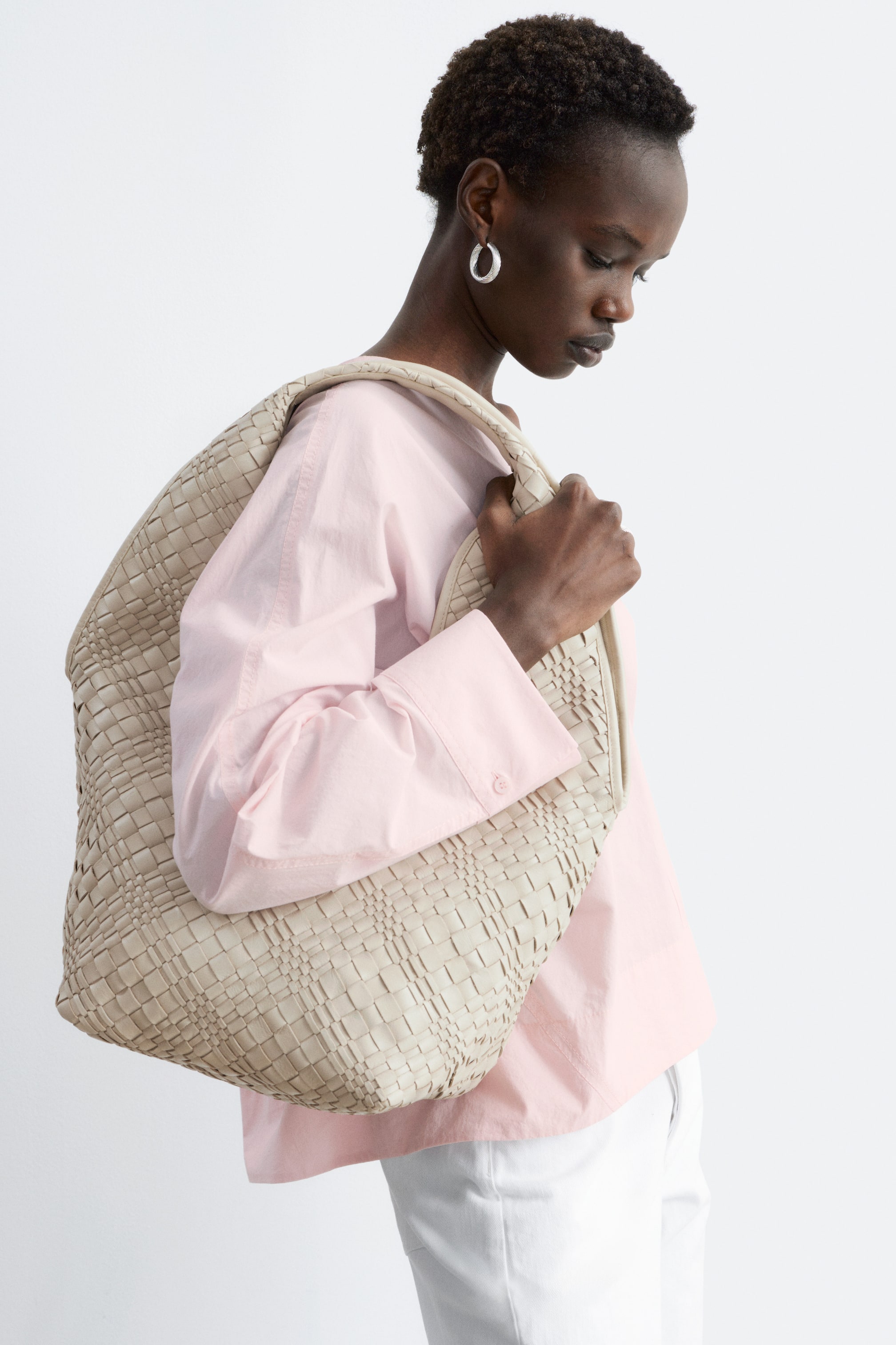 Agrandir l'image: Un grand sac tissé de couleur beige clair est porté sur l'épaule droite, légèrement incliné vers l'avant. Sa face avant et son côté présentent un motif de tissage en panier détaillé, formant une silhouette souple.