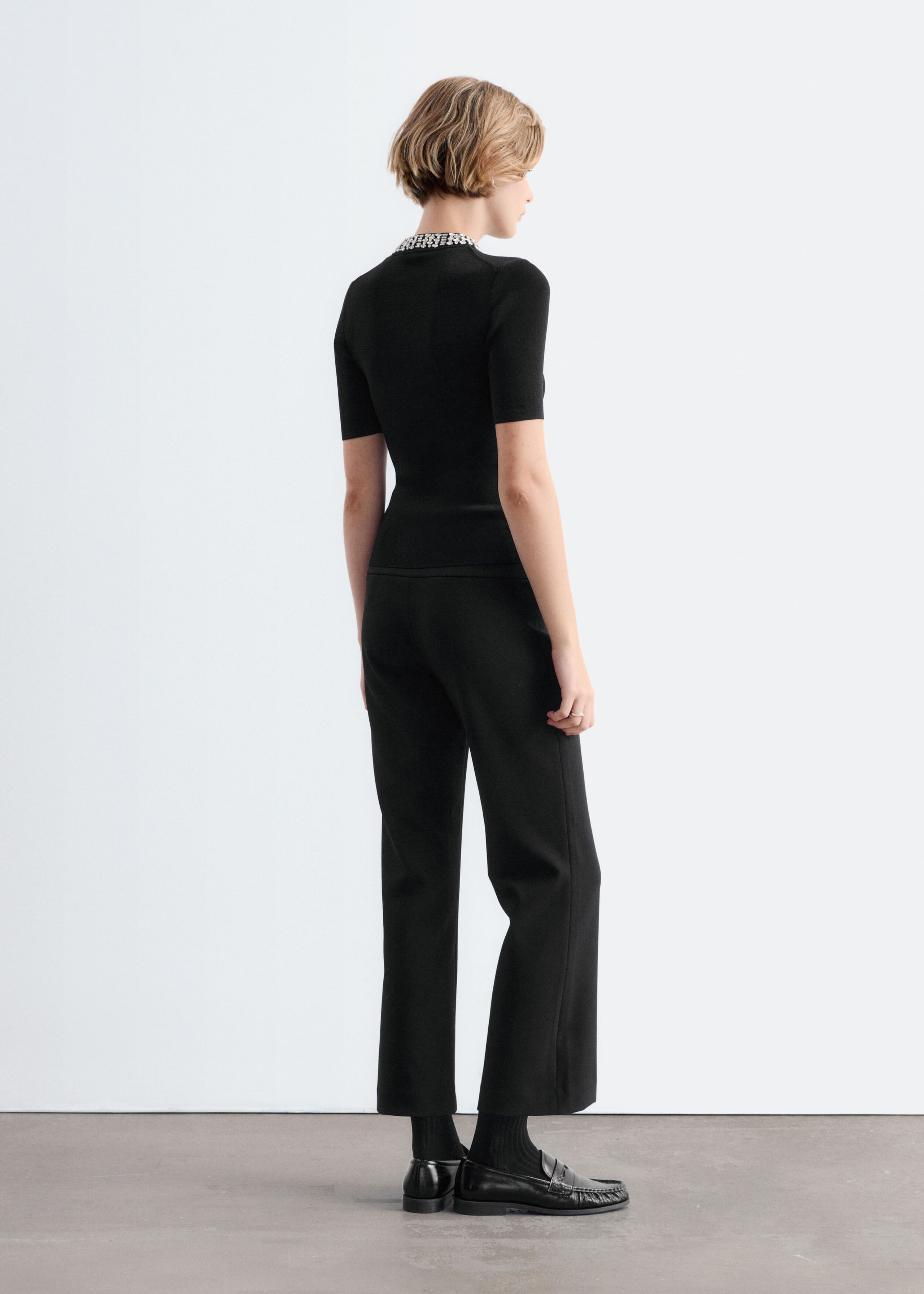 A black fitted short-sleeve top featuring a mock neck embellished with rhinestone beads is worn by the model, shown from a three-quarter rear view. The top is styled with matching black straight-leg trousers and black loafers.