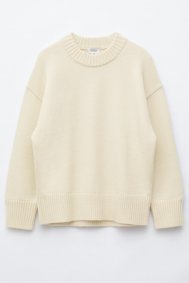 Oversizowy sweter z weÅny w kolorze kremowym przedstawiono od frontu, z obniżonÄ
liniÄ
ramion i grubym, prÄ
żkowanym okrÄ
gÅym dekoltem. Sweter ma szerokie, prÄ
żkowane mankiety oraz dopasowany szeroki, prÄ
żkowany dóÅ, które podkreÅlajÄ
jego swobodny, luźny krój. - 1