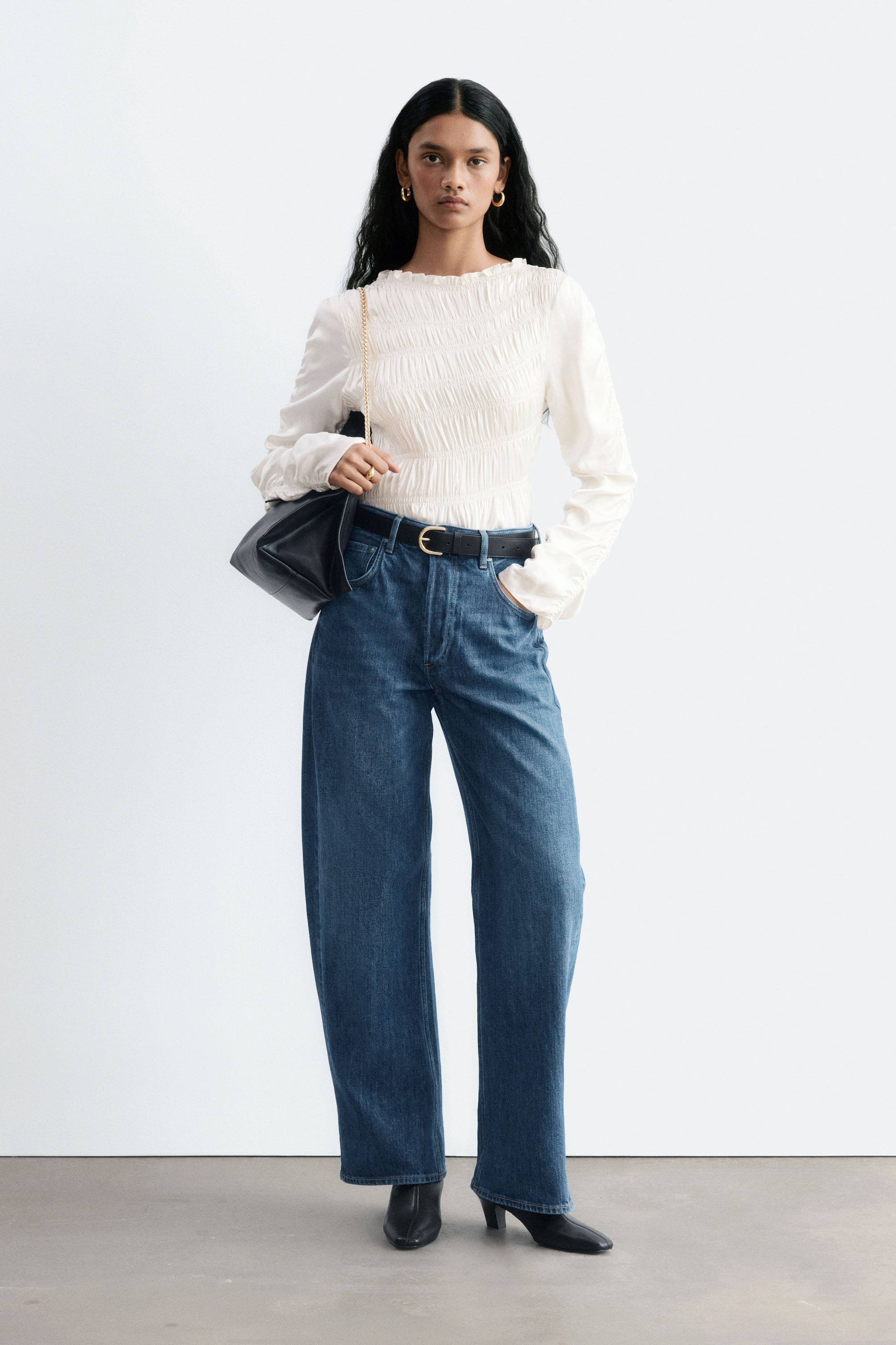 Agrandir l'image: Un jean en denim bleu à jambe large et de forme barrel est porté par le mannequin, qui se tient debout avec une main dans la poche avant. Ce jean taille mi-haute est associé à un top blanc smocké rentré à l’intérieur, une ceinture en cuir noir et des bottes noires.