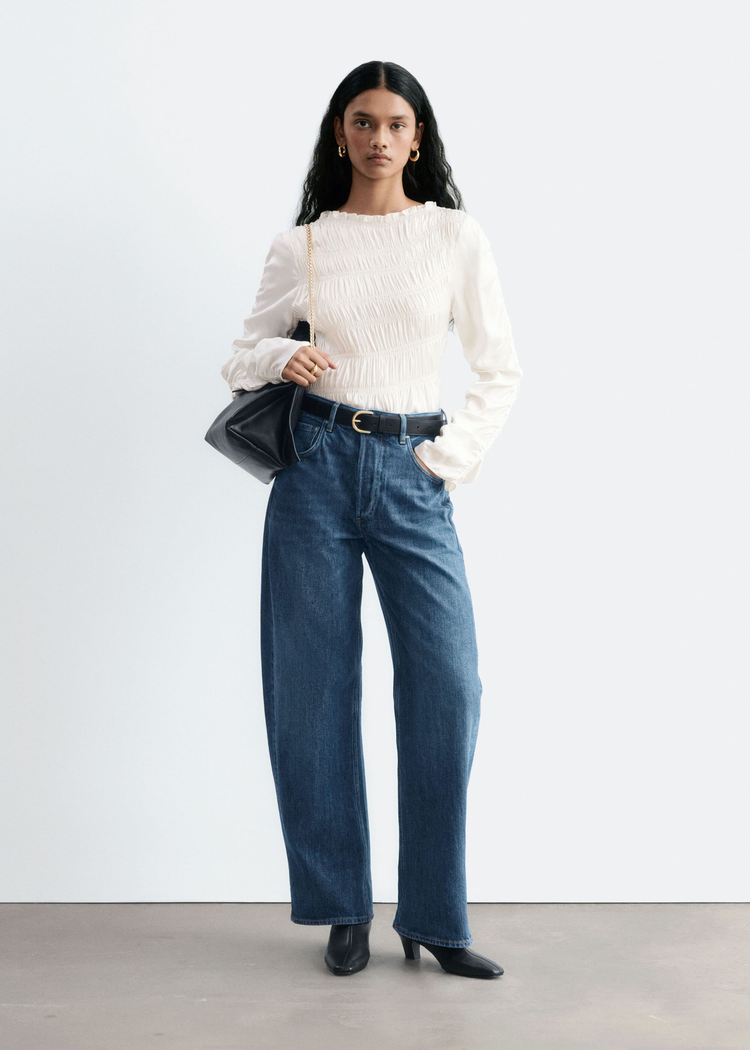 Blue denim jeans with a wide, barrel-shaped leg are worn by the model, who is standing with one hand in the front pocket. The mid-rise jeans are styled with a tucked-in white smocked top, a black leather belt, and black boots.